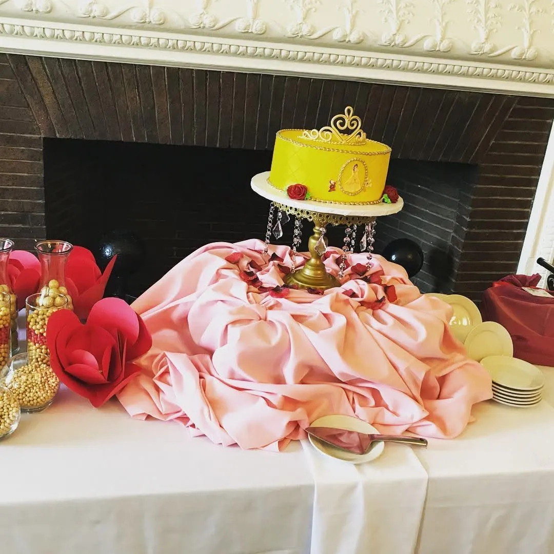 Yellow cake with decorative topper on a table with pink fabric, surrounded by red and gold decorations, in front of a fireplace.