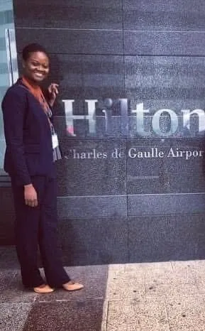 Person standing next to Hilton sign at Charles de Gaulle Airport, smiling, wearing dark suit, holding a bag, indoors.