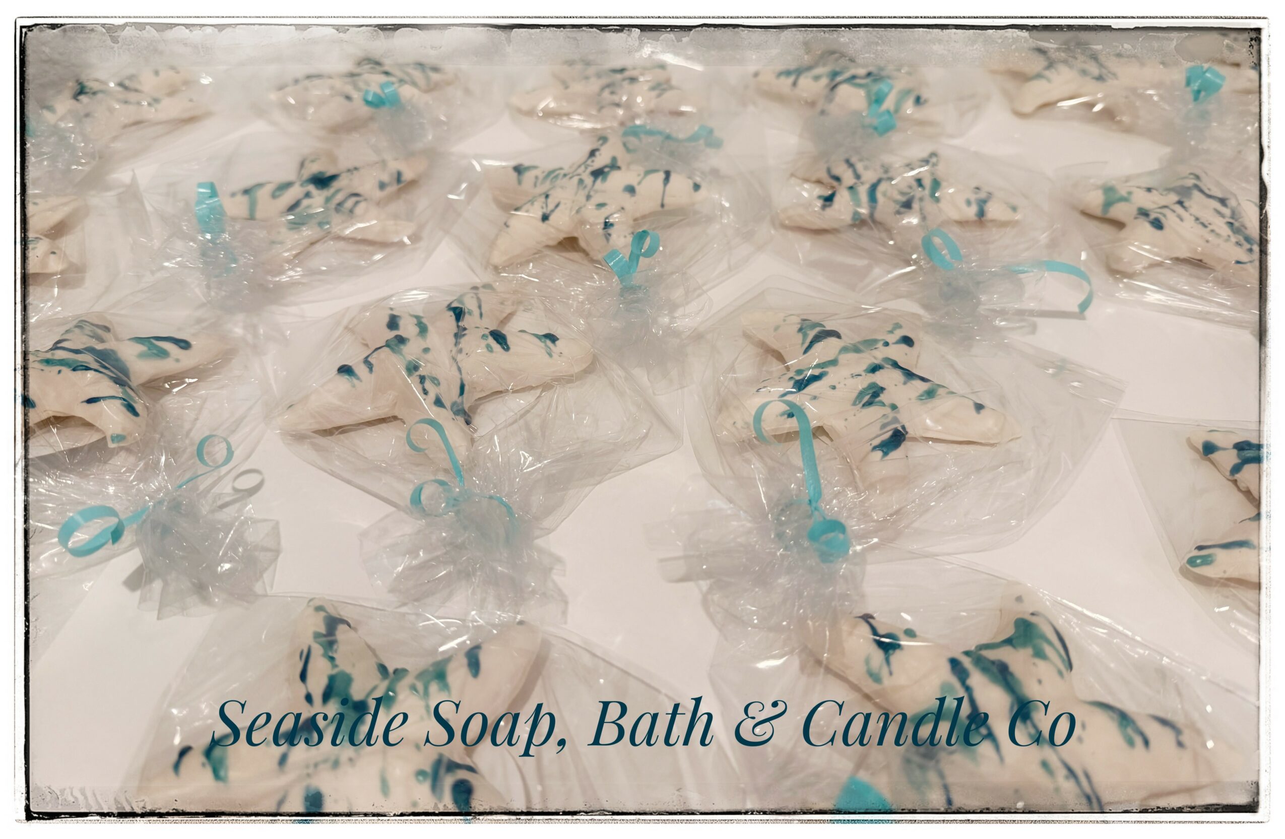Multiple soap bars wrapped in clear plastic with blue ribbons, arranged on a surface with text reading 'Seaside Soap, Bath & Candle Co.'