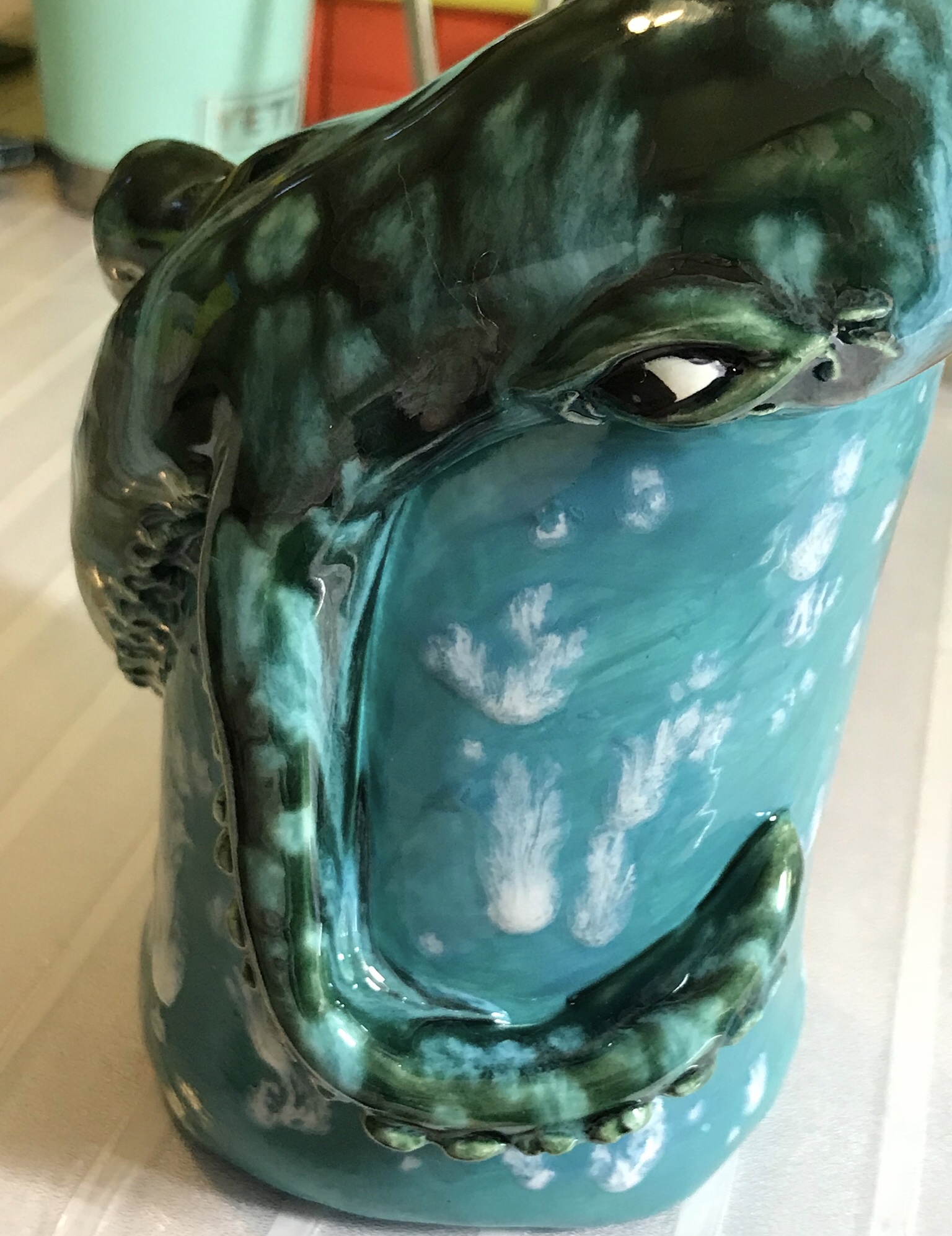 Ceramic sculpture of a face with closed eyes, painted in green and black, with a blue background and cloud-like patterns.