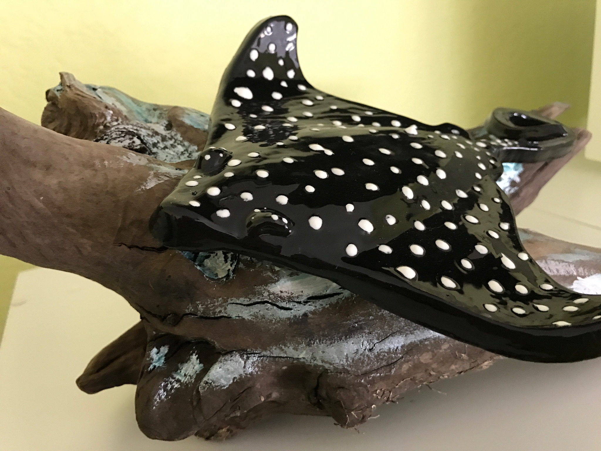 Polka-dotted black and white stingray sculpture on a piece of driftwood, with a yellow wall in the background.