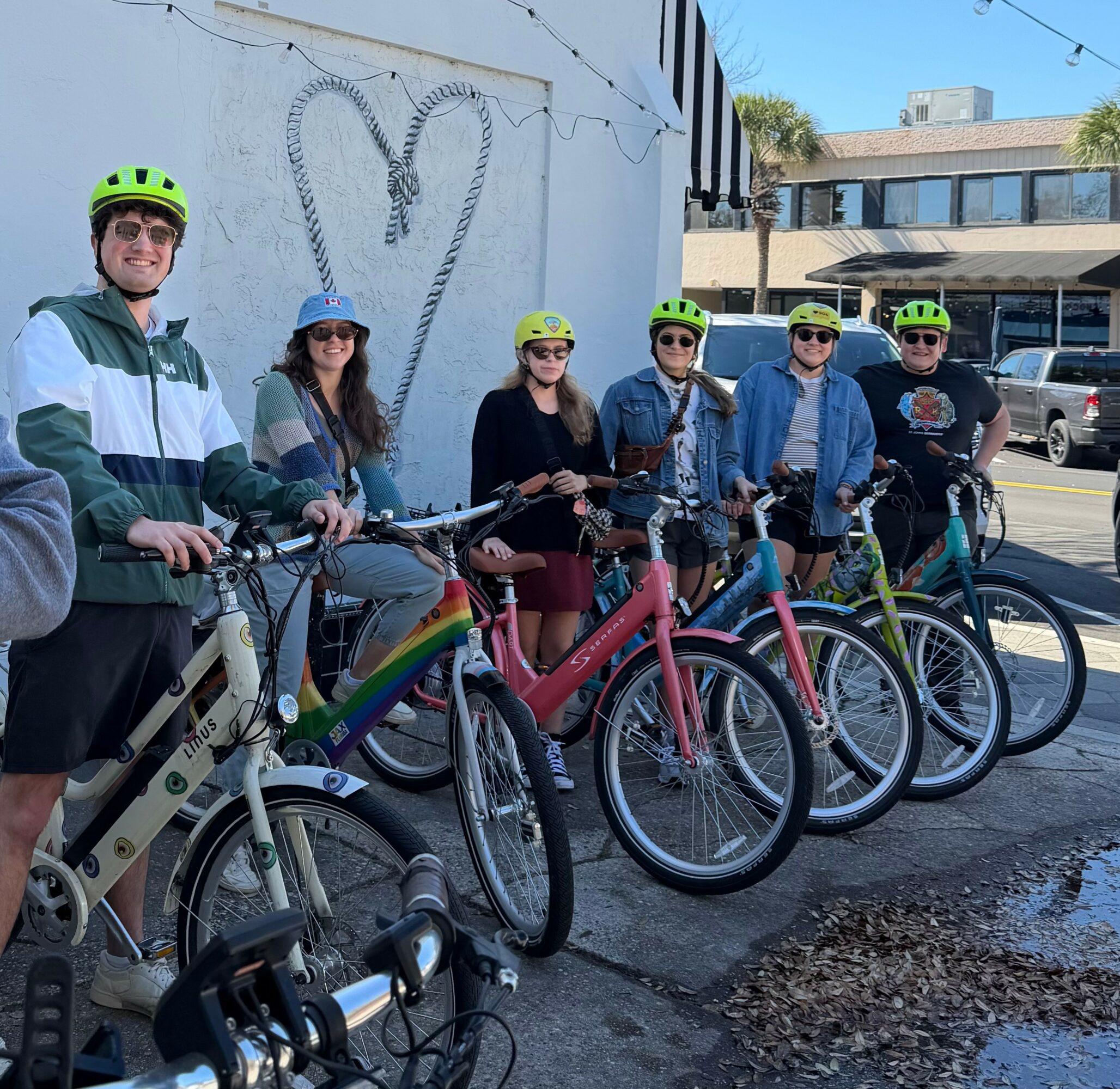 Group of seven people with bikes standing on sidewalk in front of a wall with a heart-shaped decoration, sunny day.