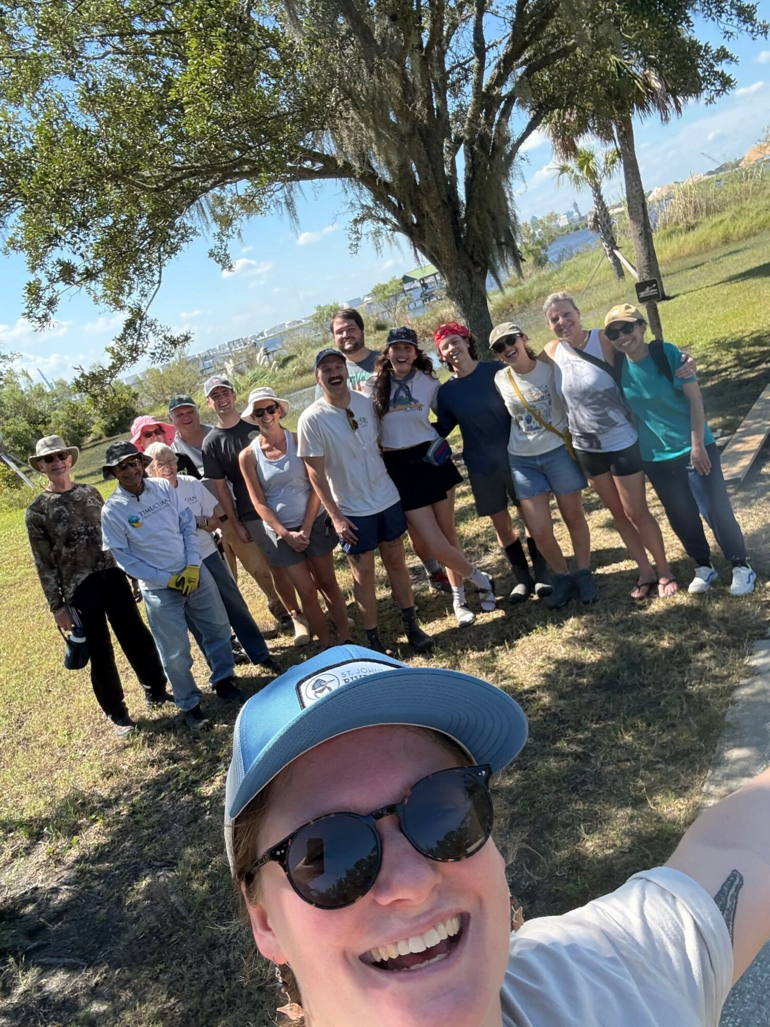 Person wearing sunglasses and a helmet taking a selfie with a group of people outdoors near a body of water, trees, and grass.