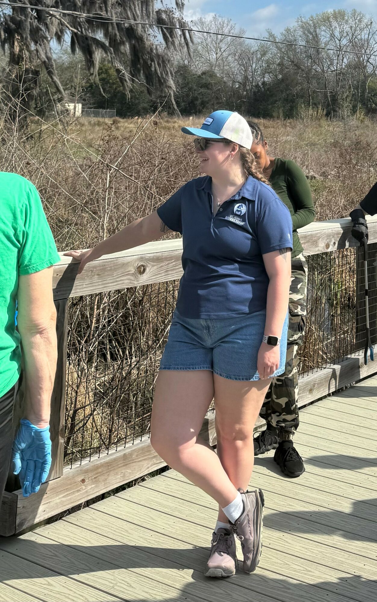 Woman in navy shirt, shorts, and cap standing on a wooden bridge, with trees and bushes in background.