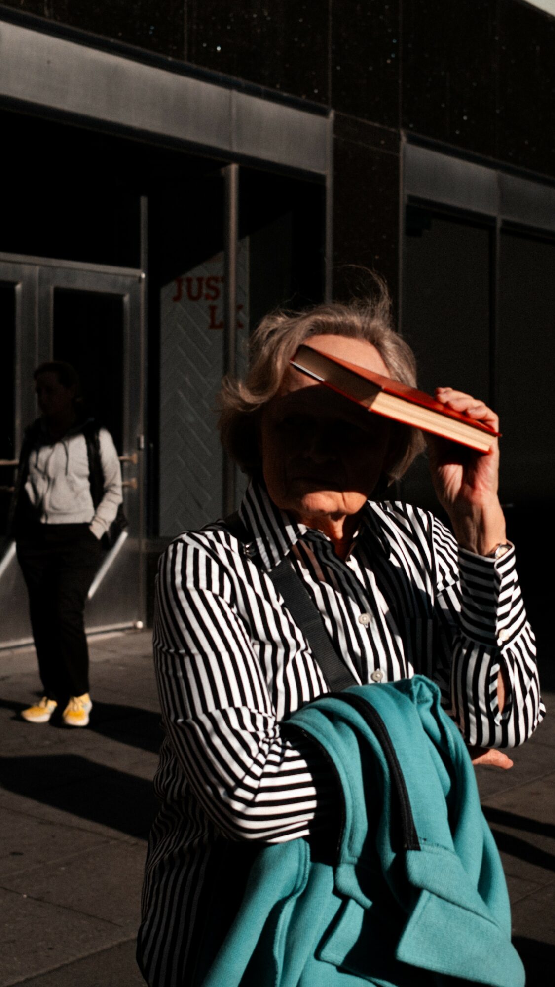 Person with striped shirt holding a book over their face, standing outside a building, with another person in the background.