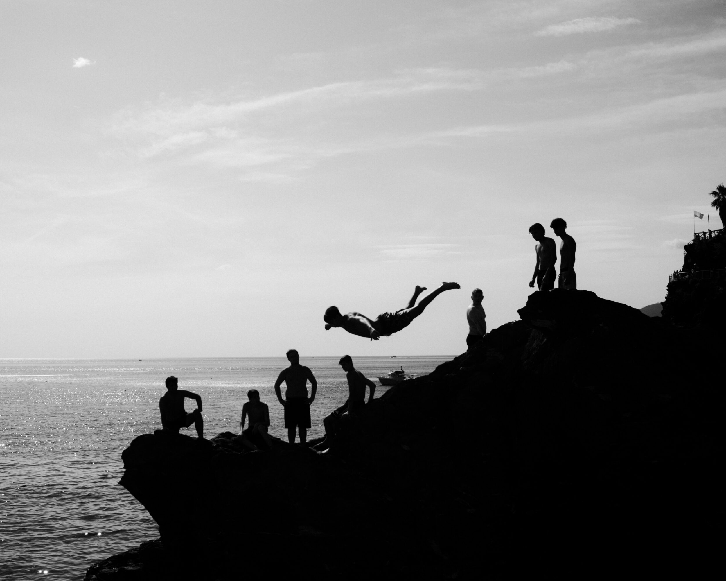 Silhouettes of people on rocks by the sea, one person mid-air diving into water, cloudy sky, black and white.