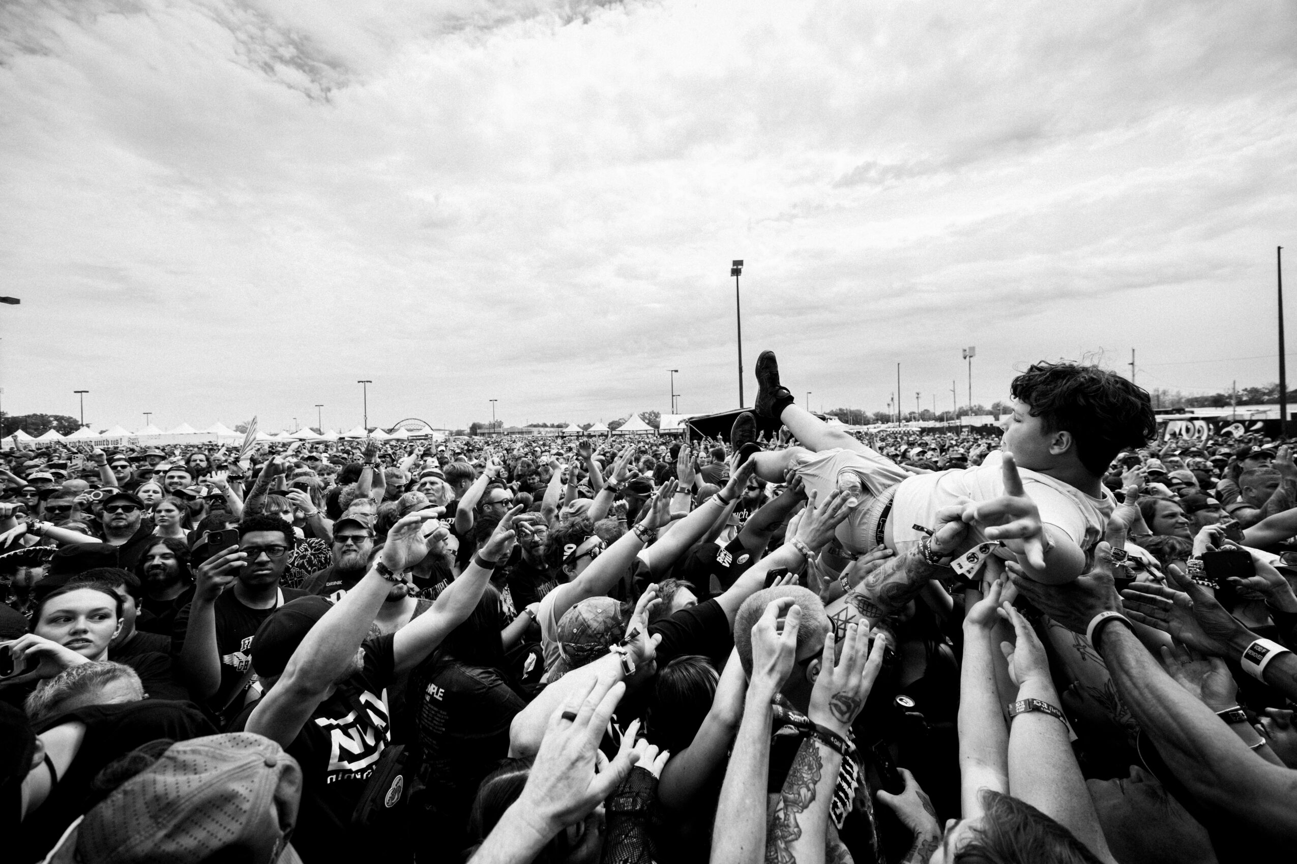 Large crowd of people reaching out towards a person lying on the ground outdoors, with a cloudy sky and distant structures.