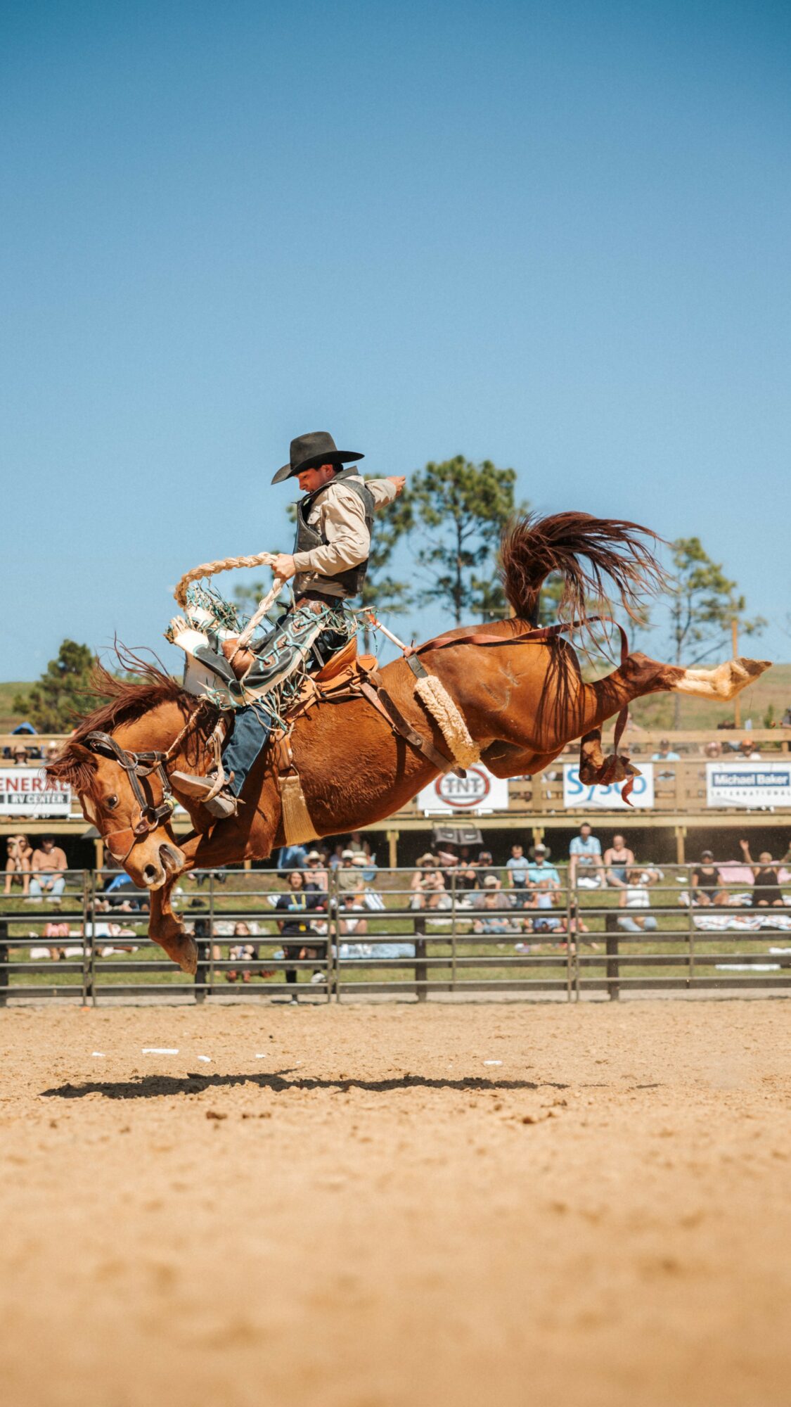 Cowboy riding a bucking horse during a rodeo event, with spectators in the background and clear blue sky.