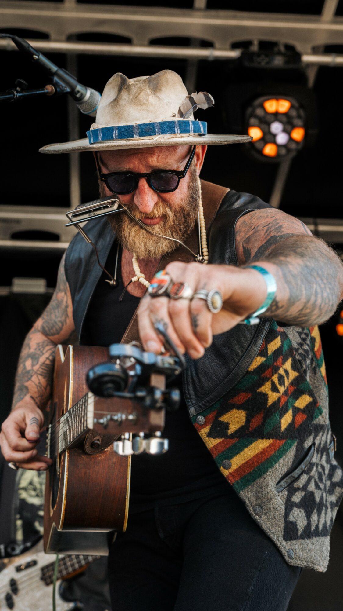 Man with tattoos, sunglasses, and a hat playing guitar outdoors, with stage lights in background.