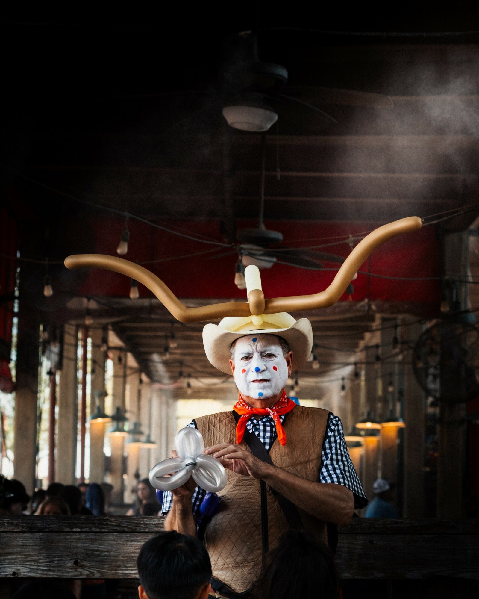 Person dressed as a clown with a hat and painted face, holding a balloon, in a dimly lit indoor setting.