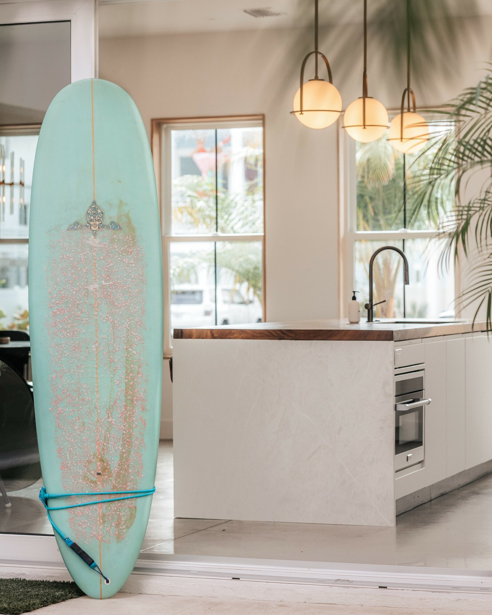 Surfboard standing upright in a bright kitchen with large windows, pendant lights, and a plant, near a white counter and sink.