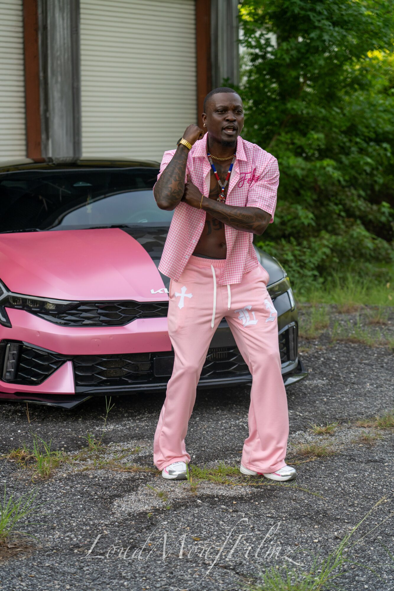 Man in pink outfit standing in front of a pink car outdoors, with trees and a building in the background.
