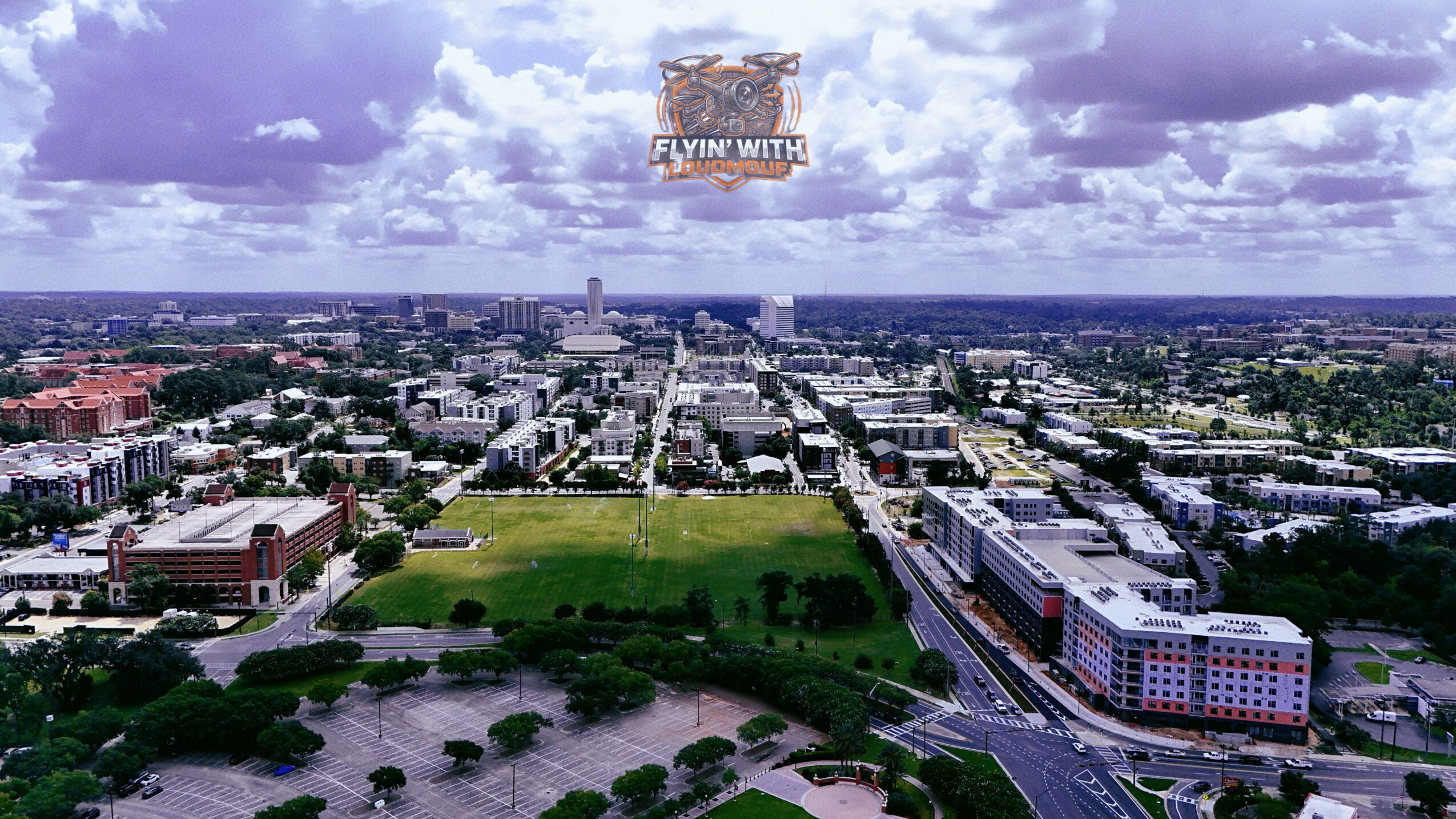 Aerial view of a city with a large green park, buildings, and roads under a cloudy sky.