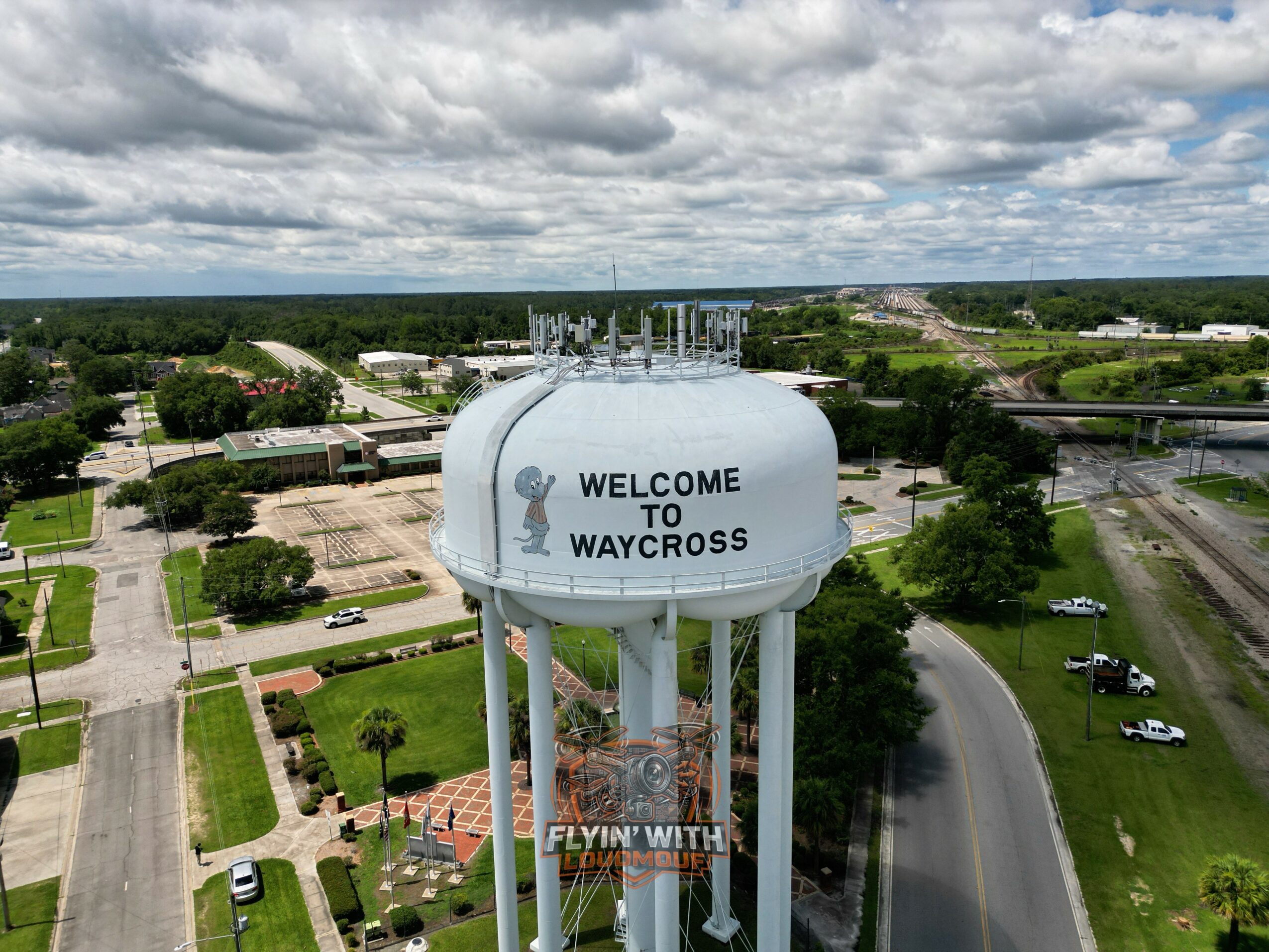 Large white water tower with 'WELCOME TO WAYCROSS' text, surrounded by trees and roads, under a cloudy sky.