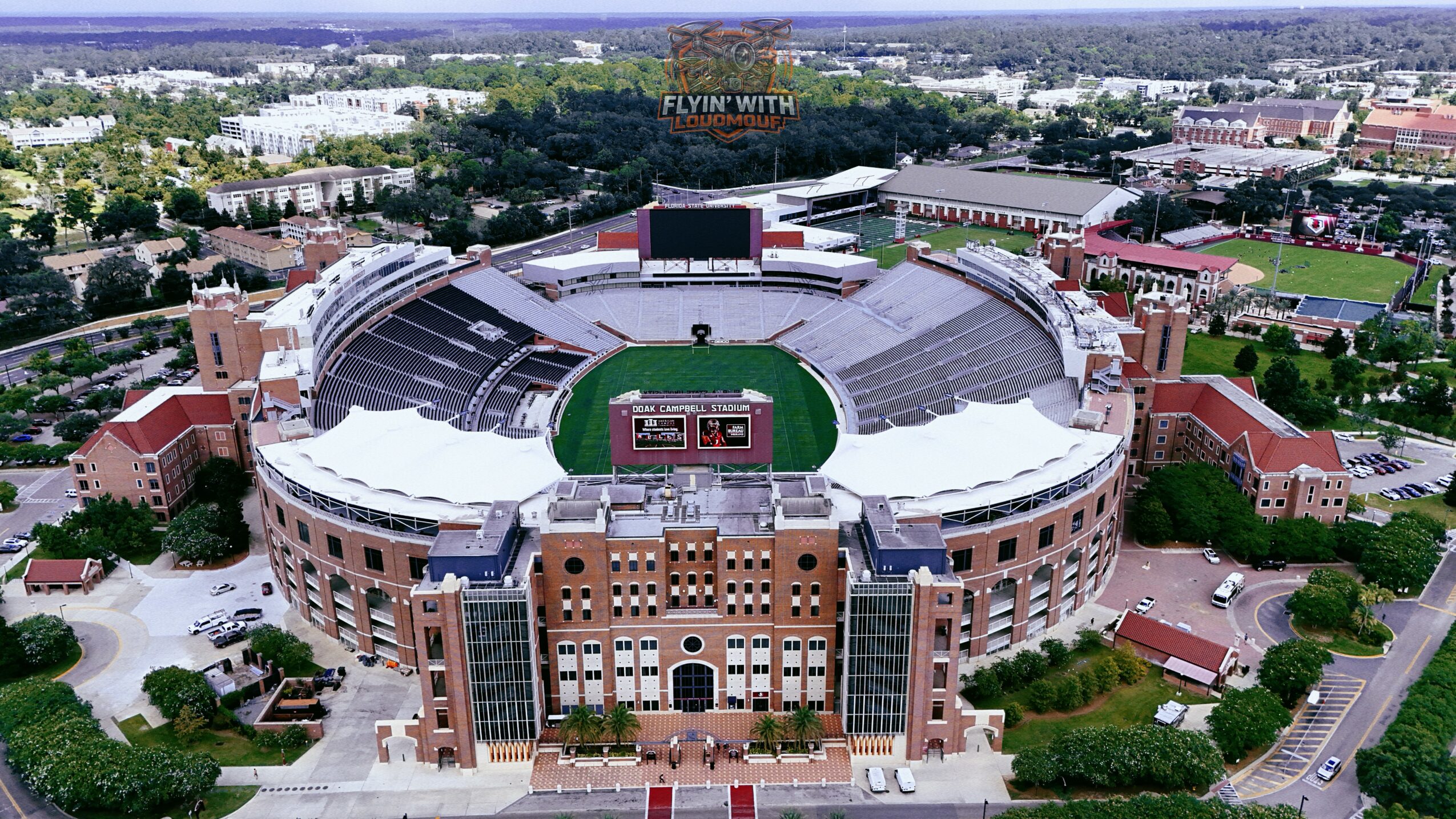 Aerial view of a large stadium with a green field, surrounded by seating and buildings, in a cityscape.