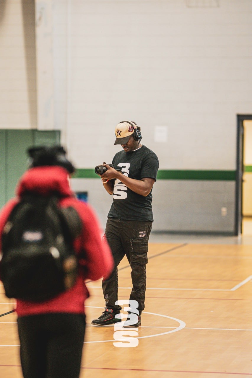 Person wearing a black shirt, black pants, and a helmet, standing in a gymnasium, looking at a device, with another person in red in foreground.