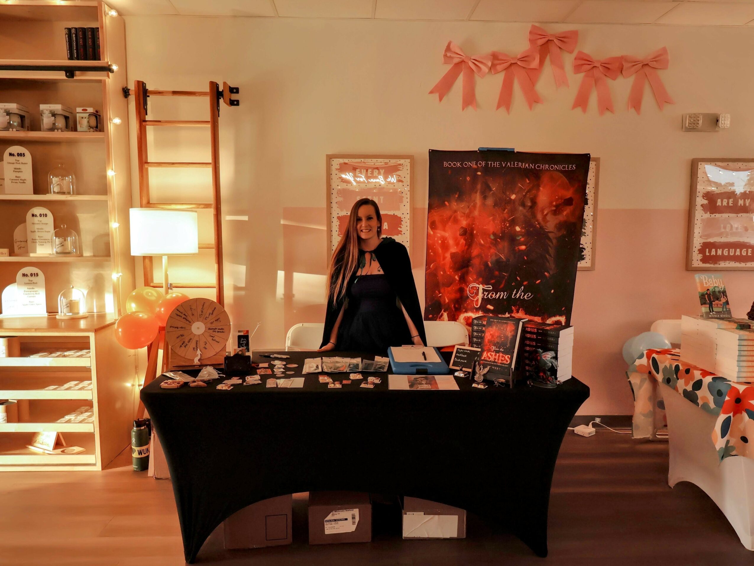 Woman standing behind a table with books and items, decorated with pink bows on the wall, in a cozy room.