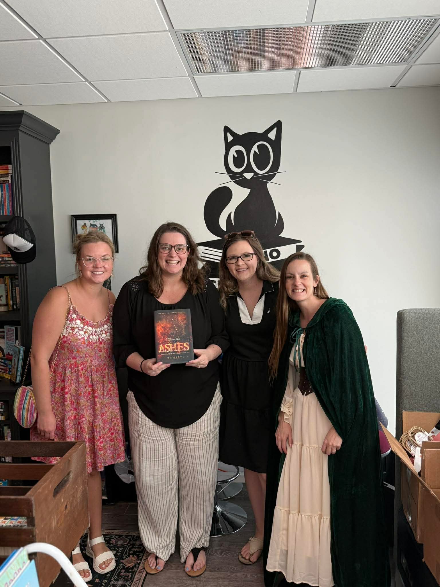 Four women standing indoors, smiling, with a black cat wall decal above them, holding a book, in a room with bookshelves.