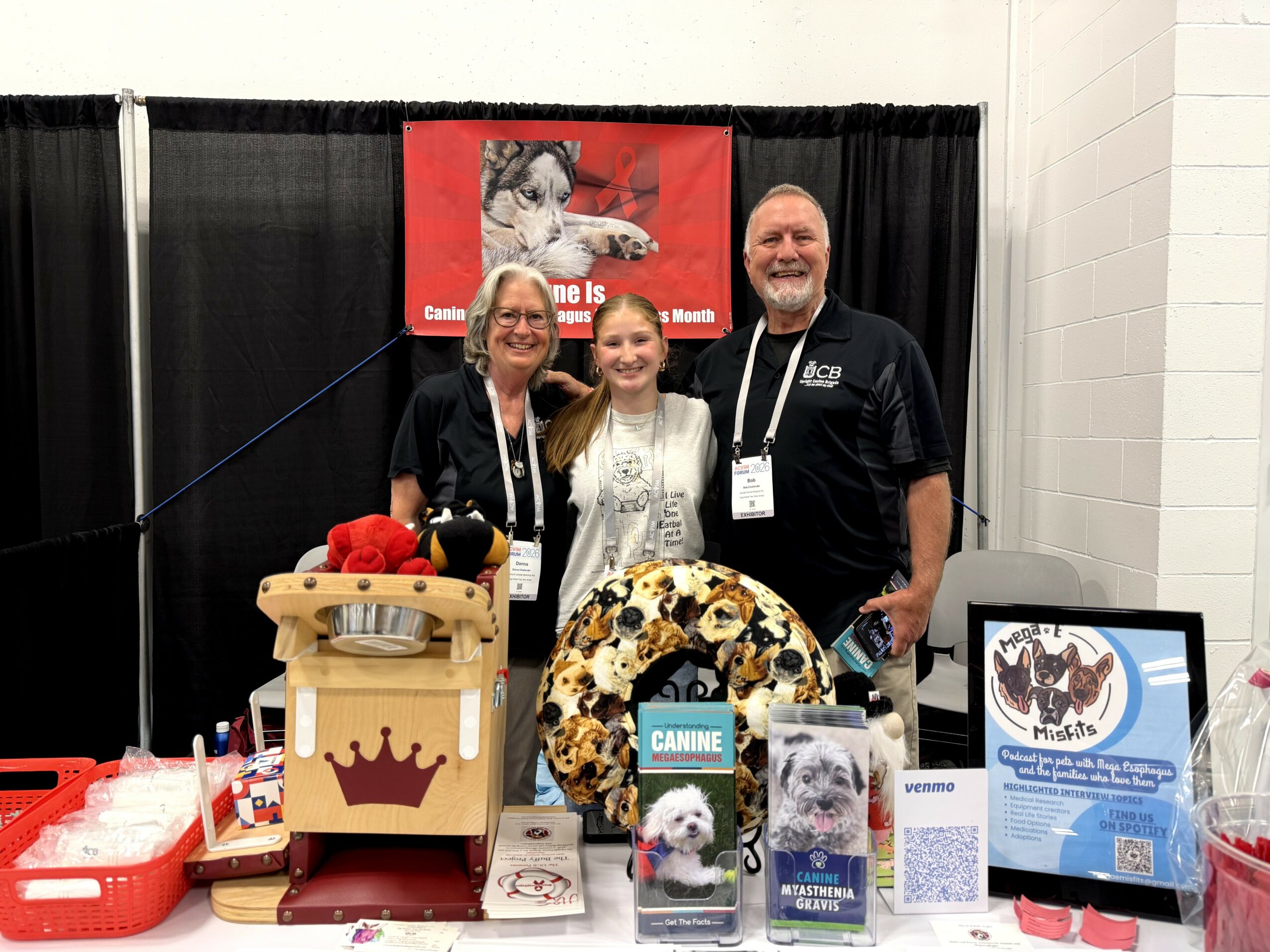 Three people stand behind a table with dog-related items, a red banner, and a black curtain background.