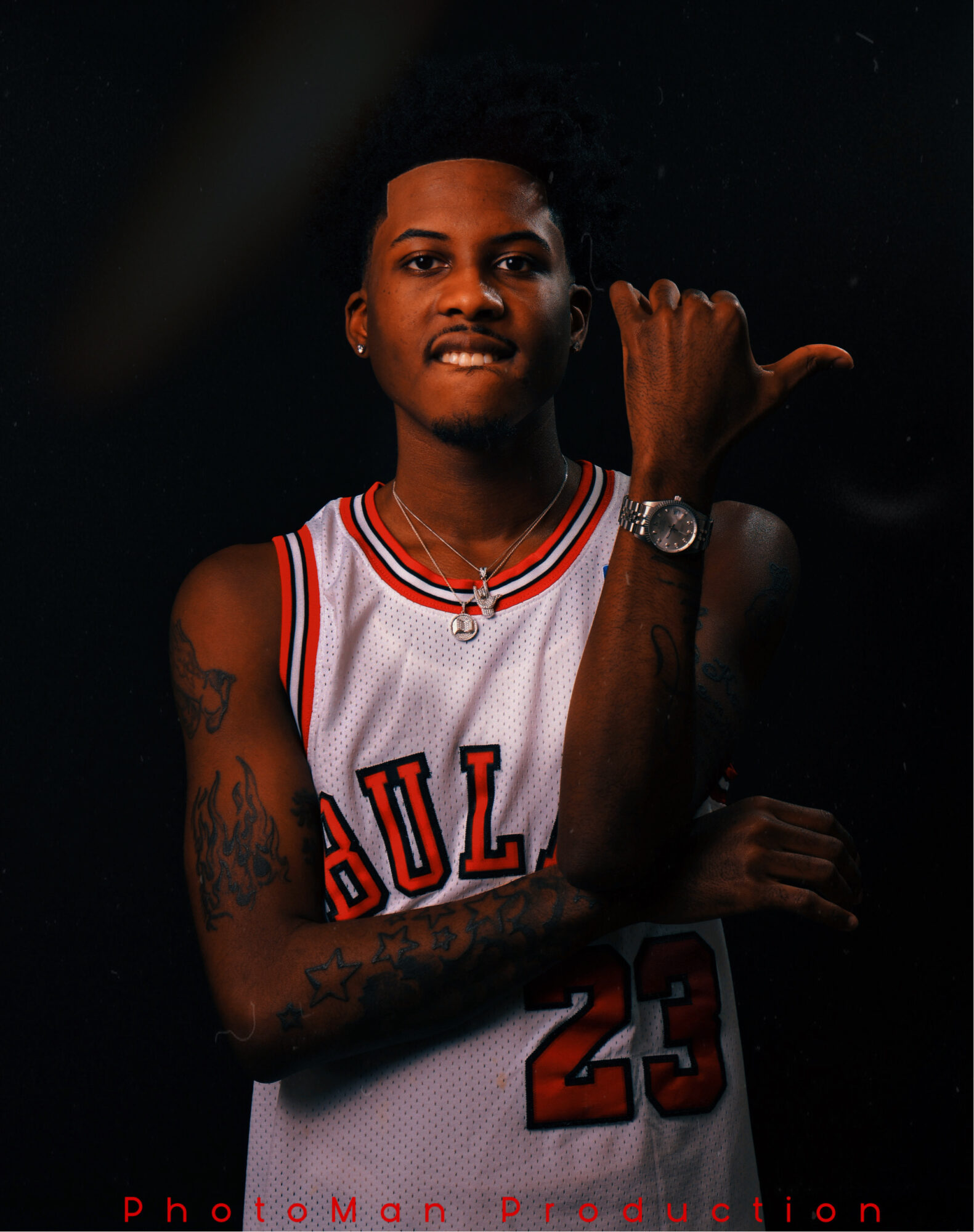Person wearing a basketball jersey with the number 23, smiling, showing a tattooed arm and a watch, making a gesture with their hand.