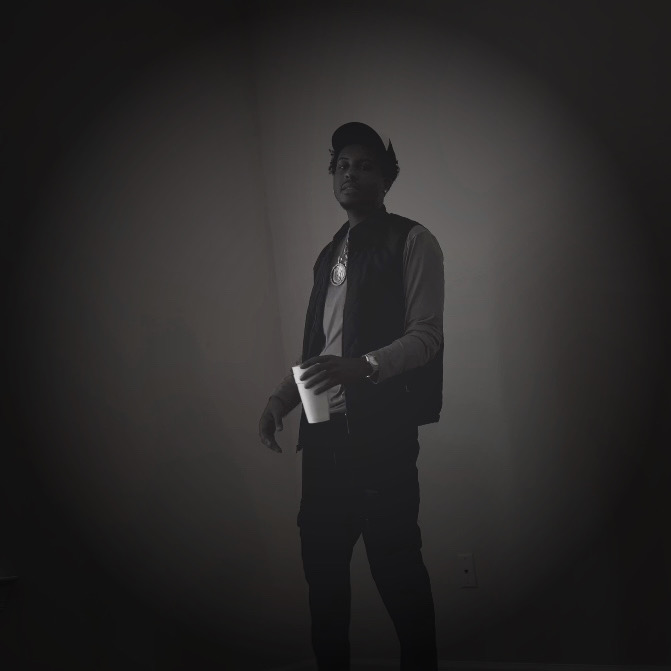 Person standing in a dark room holding a cup, wearing glasses, a cap, and a vest, with a spotlight on them.