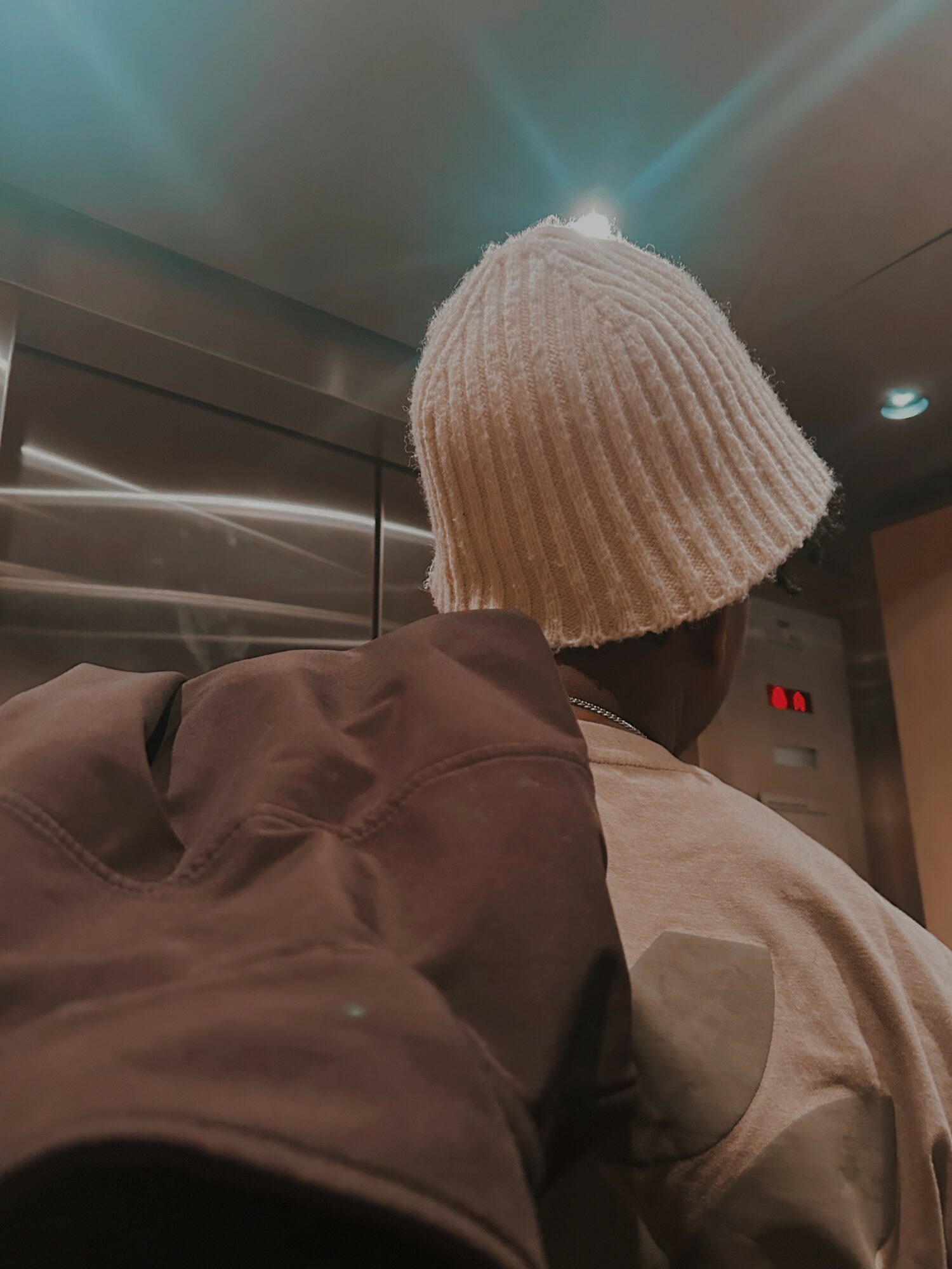 Person wearing a white knit hat and brown jacket, seen from behind in an elevator.
