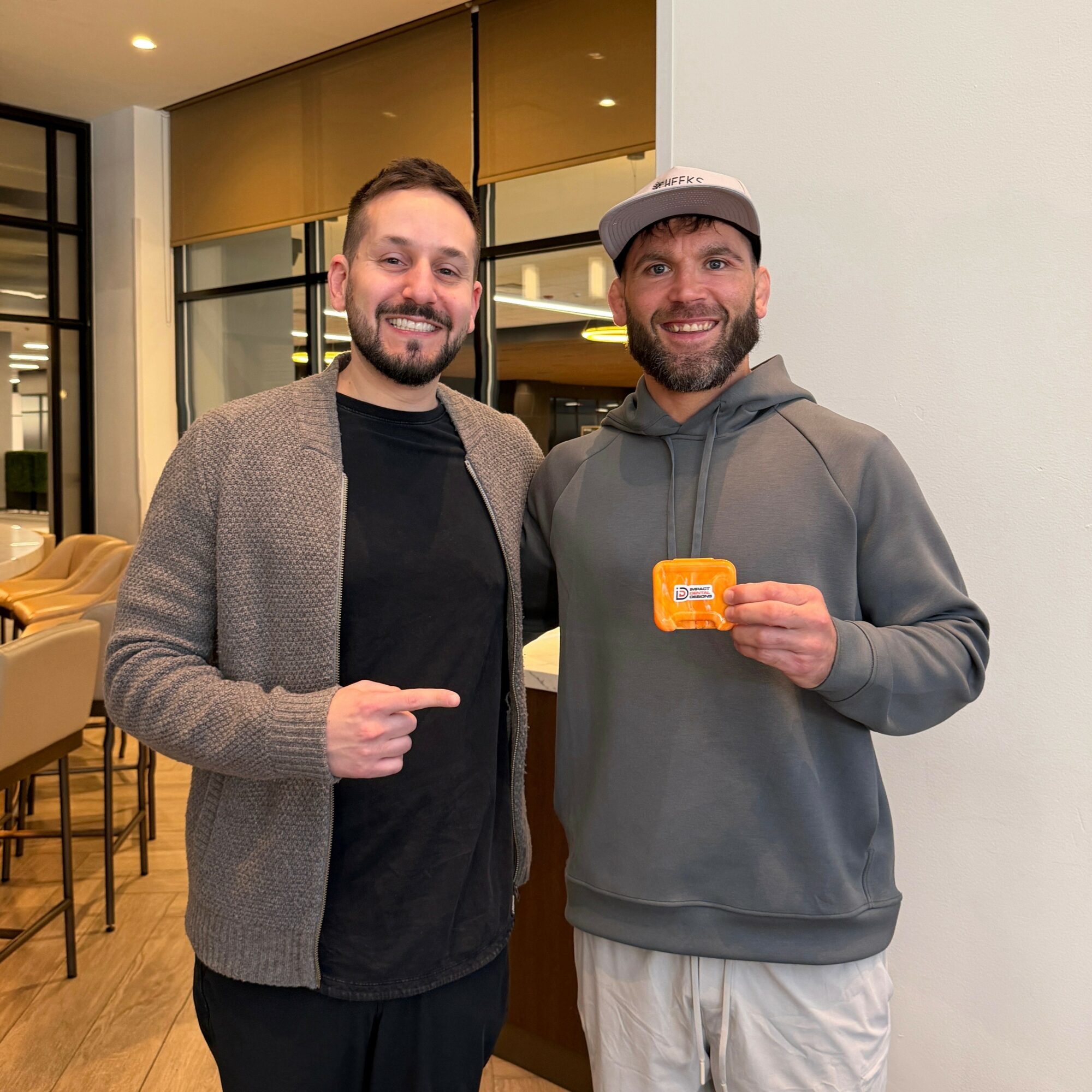 Two men smiling indoors, one pointing at the other who holds a small orange card, casual clothing, modern interior.