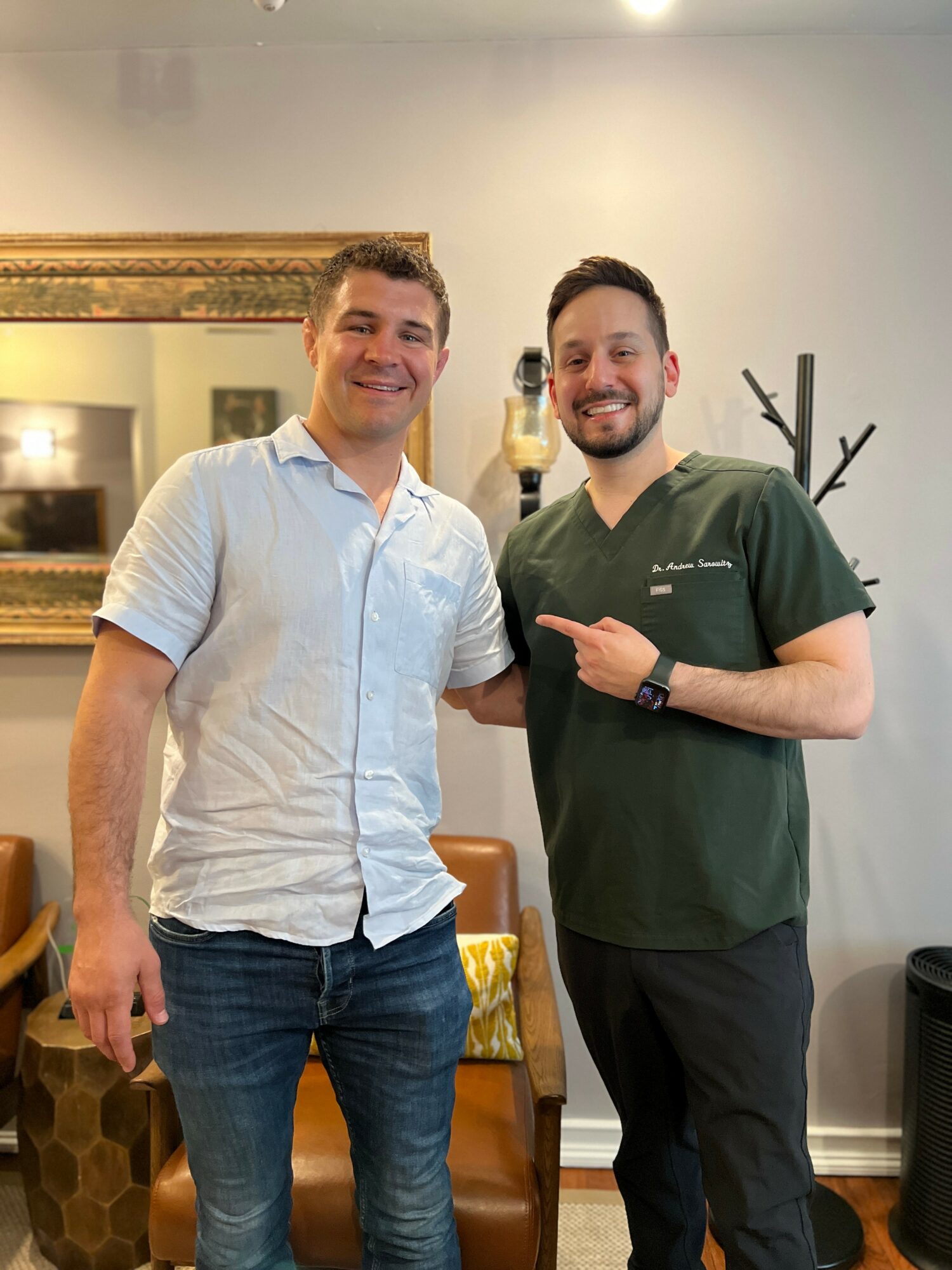 Two men smiling indoors, one in a white shirt and jeans, the other in a green medical uniform, pointing at each other.