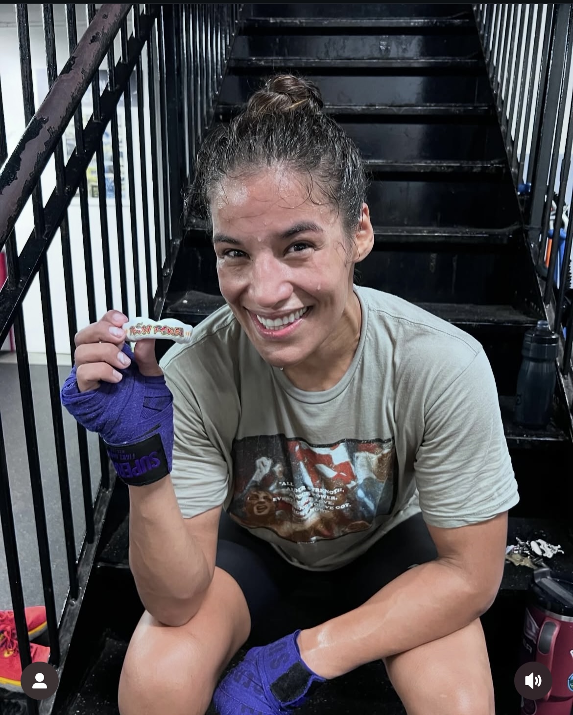 Woman smiling, sitting on stairs, holding a small object, wearing a light-colored T-shirt and purple gloves.