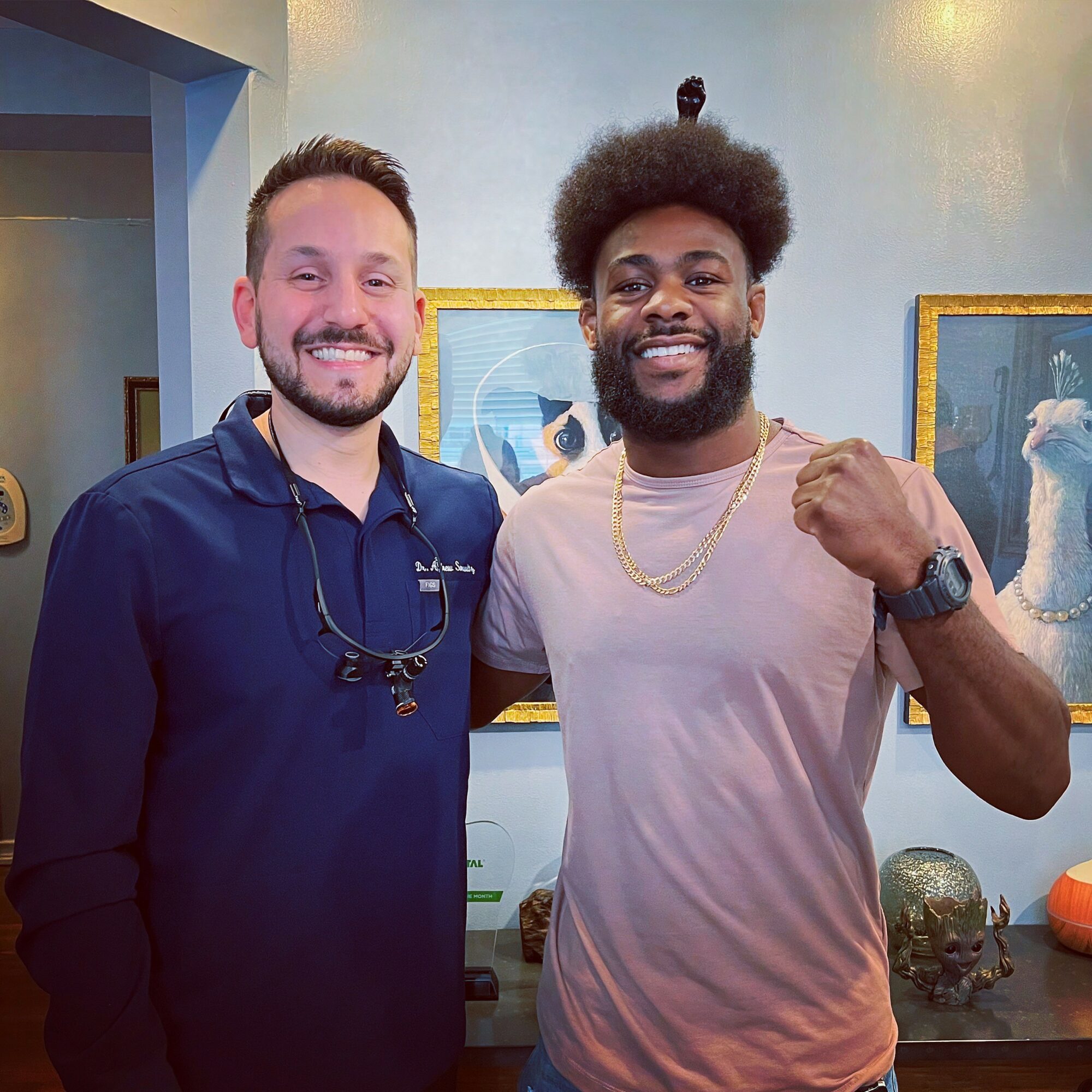 Two men smiling indoors, one with short hair and beard, the other with curly hair and beard, standing in front of paintings.