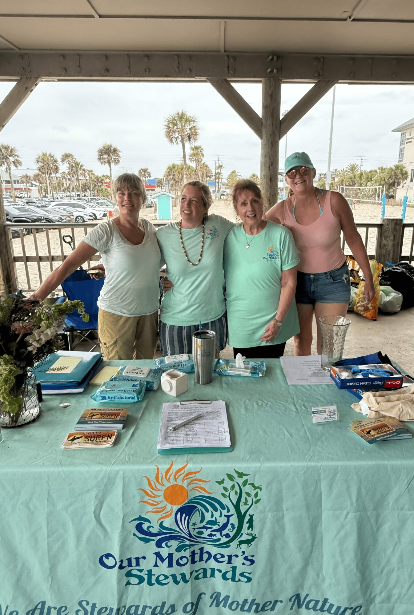 Four women stand behind a table with a banner reading 'Our Mother's Stewards' under a pavilion, with boats and palm trees in the background.