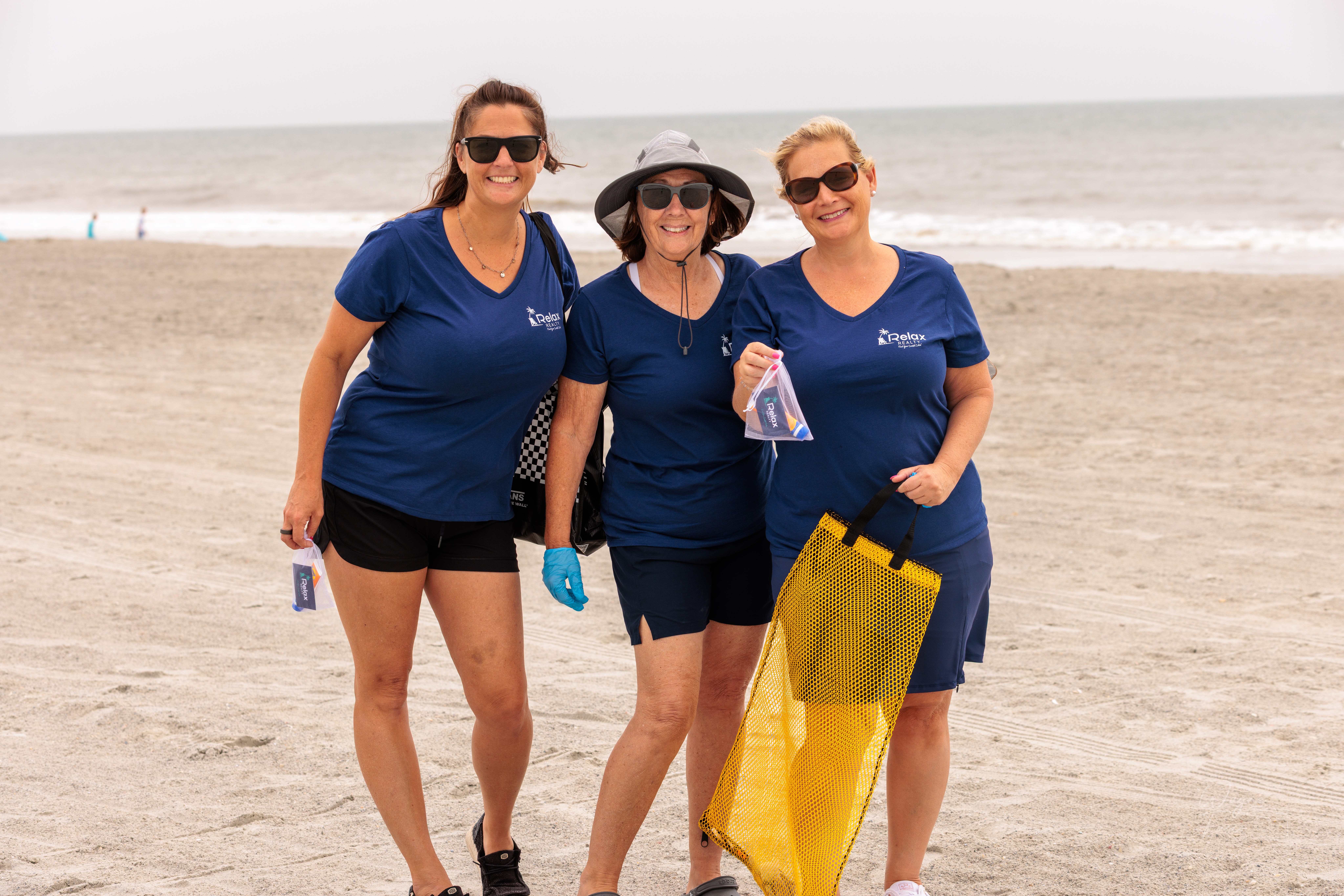 Three women in blue shirts and shorts standing on a beach, smiling, holding bags and drinks, with the ocean in the background.