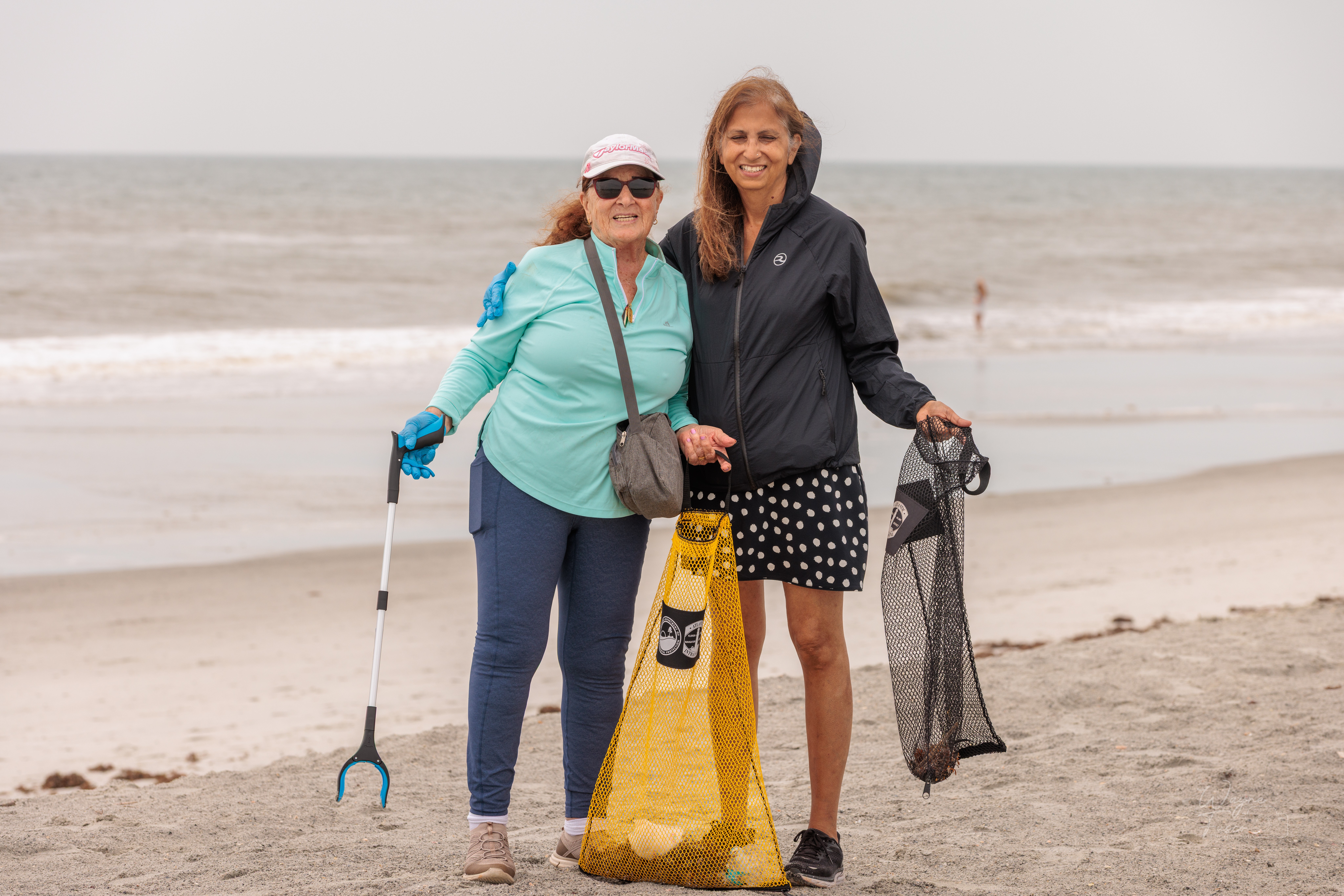 Two women stand on a beach holding a yellow trash bag and a fishing net, smiling at the camera.