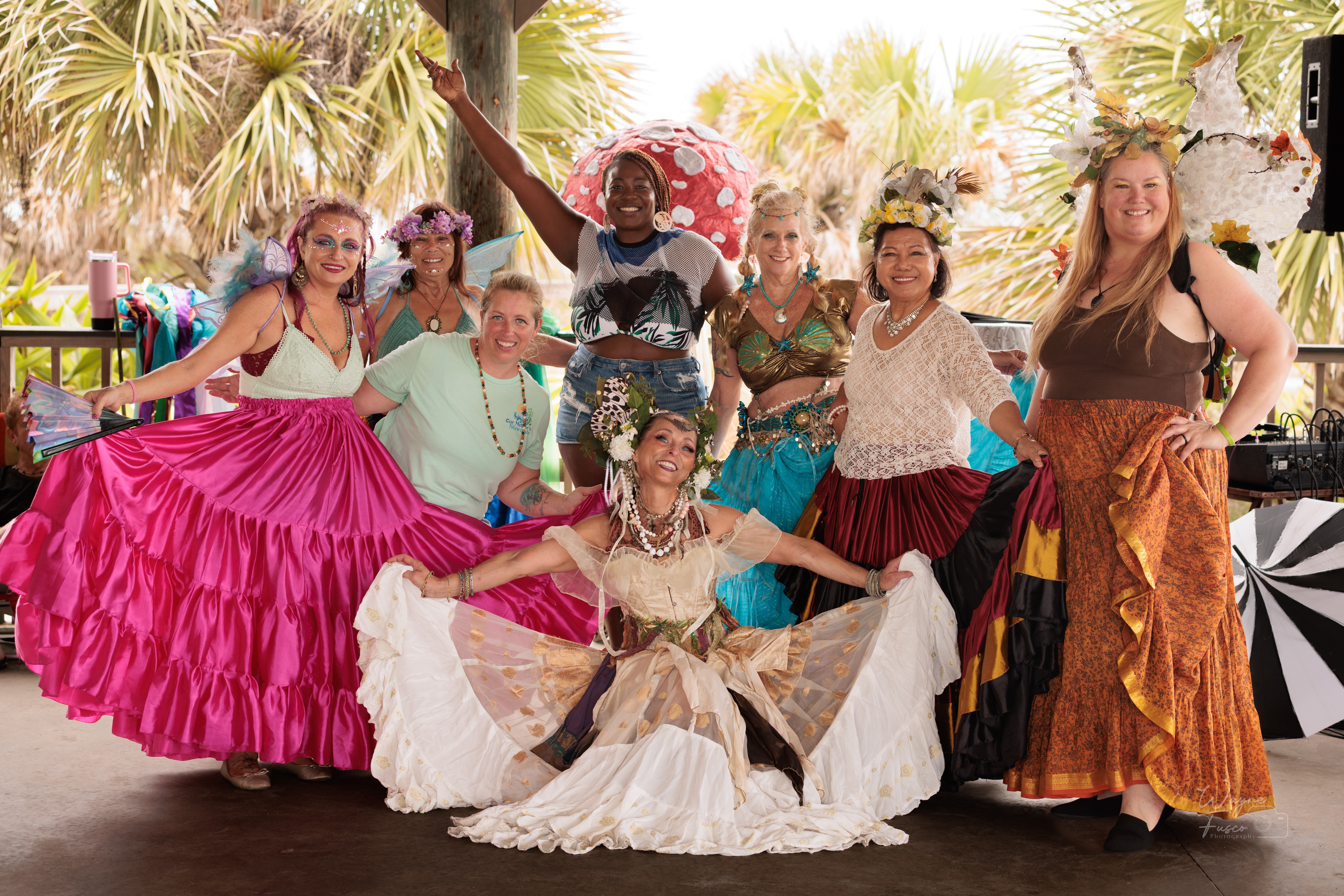 Group of women in colorful, traditional dresses smiling outdoors with palm trees in background.