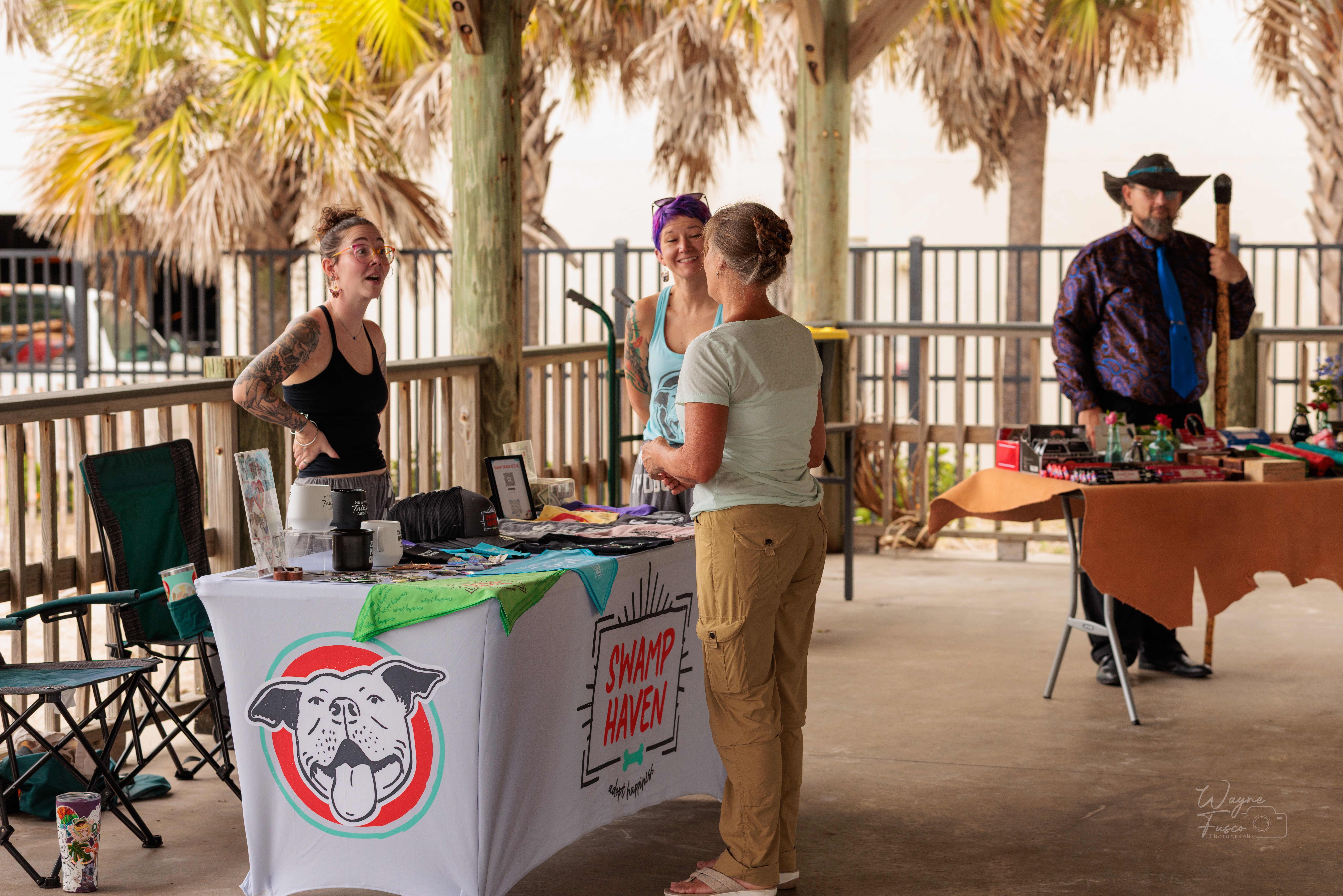 Four people at an outdoor market stall with a colorful banner, under palm trees, engaging in conversation, with tables and chairs nearby.