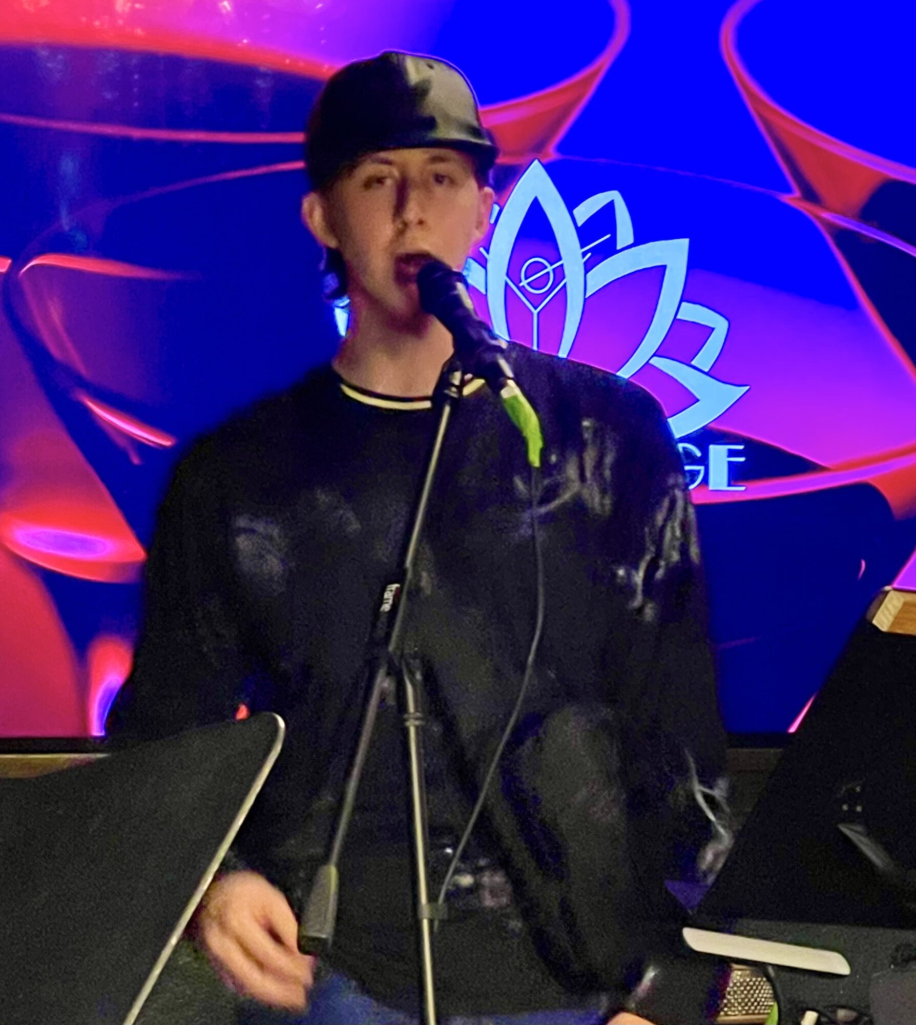 Young person singing into a microphone on stage with a colorful background, wearing a black cap and jacket.