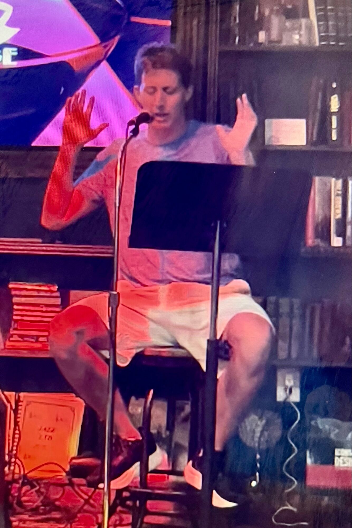 Person sitting on a chair with a microphone, wearing a pink shirt and shorts, gesturing with hands, in a room with bookshelves.