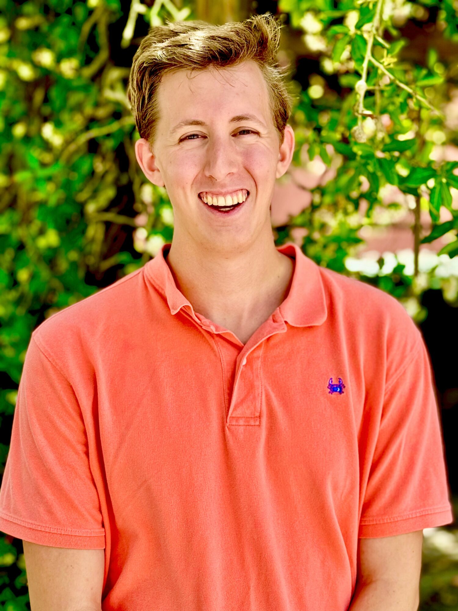Young person with short hair smiling outdoors, wearing a coral polo shirt, surrounded by green foliage.