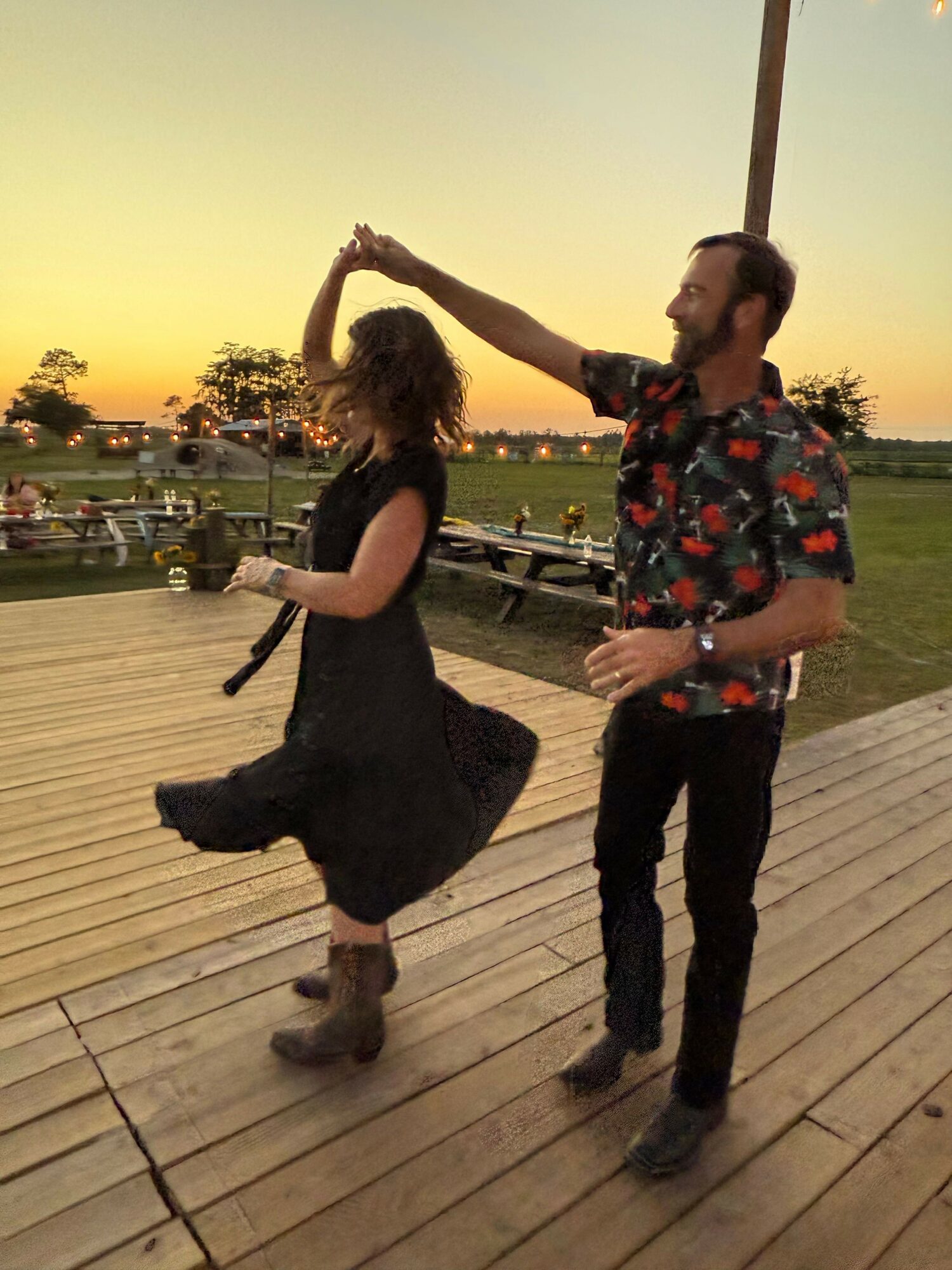 A woman is bending down to touch a man's head on a wooden deck during sunset, with a cityscape in the background.