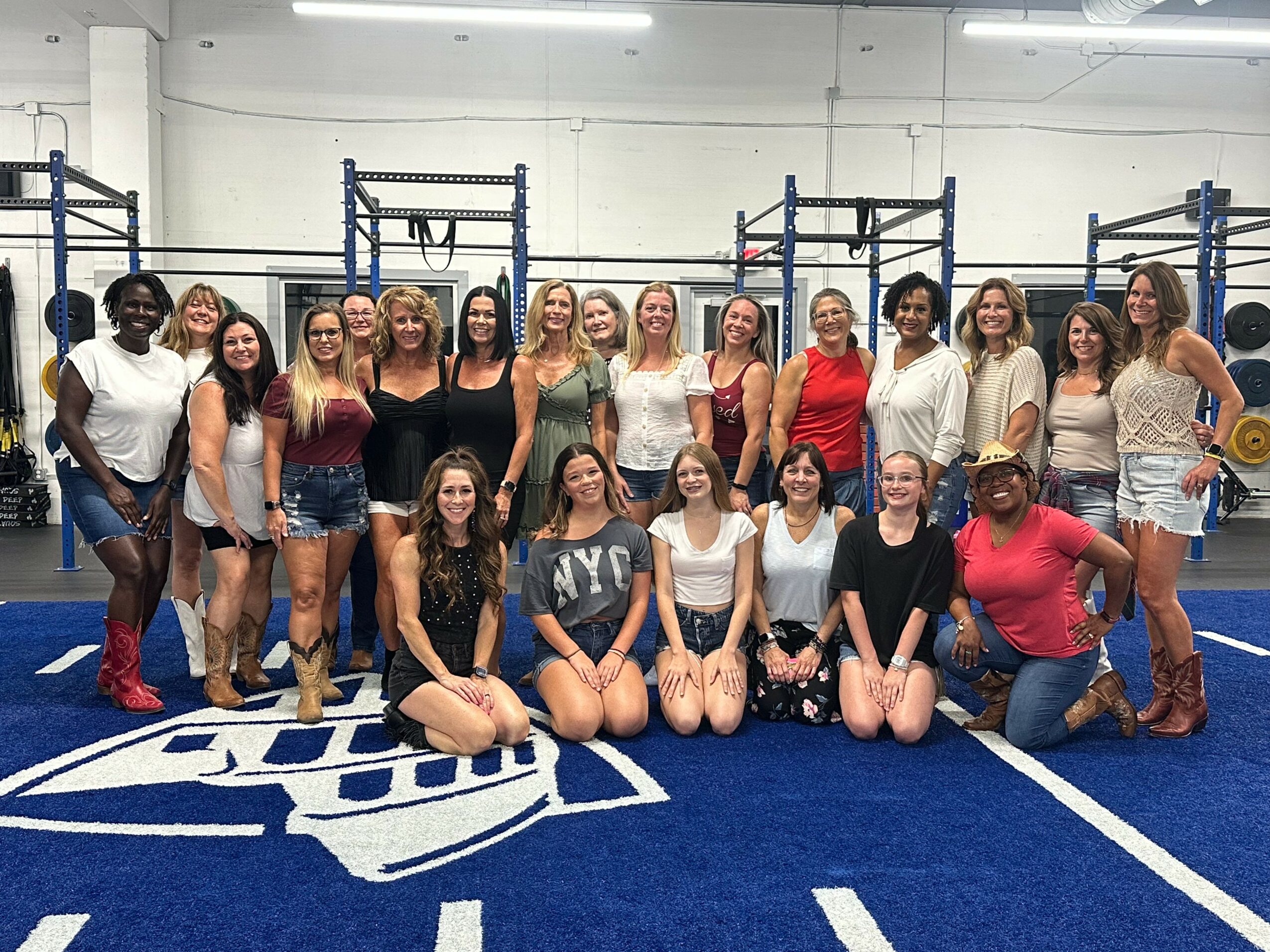 Group of women and girls posing in a gym with blue flooring and equipment, some standing and some kneeling, smiling.