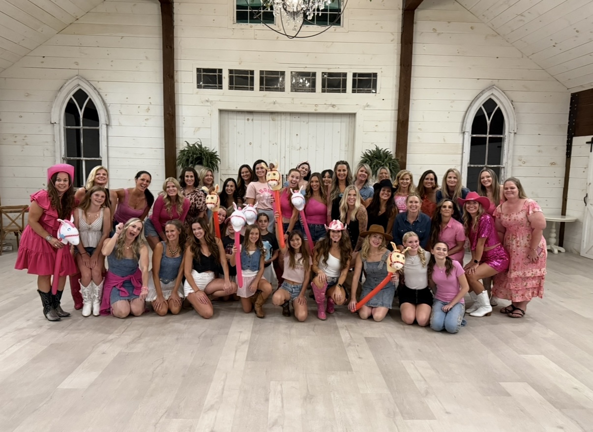 Group of women and girls gathered in a church hall, smiling and posing for a photo, some holding pink bags.