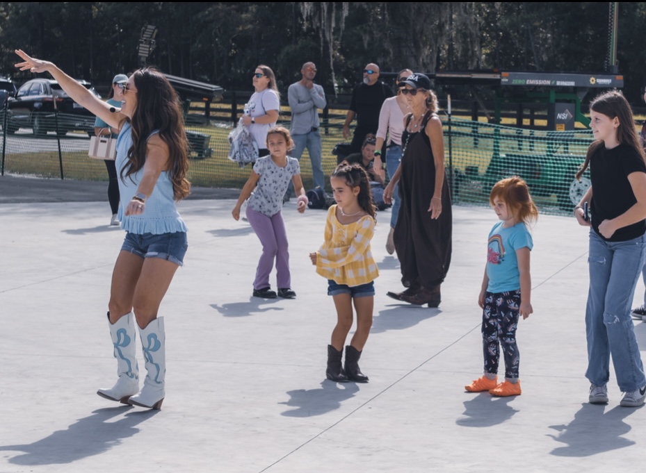 Group of children and adults outdoors, some children dancing or playing, adults watching, sunny day, park setting.