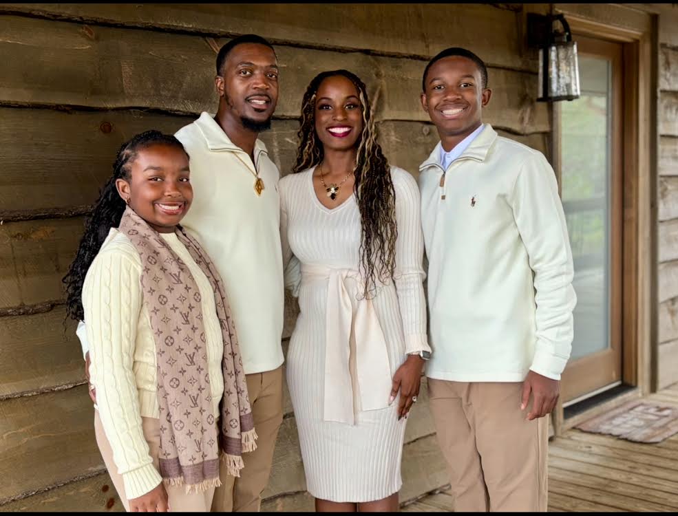 Four people stand together indoors, smiling, with a wooden wall background and a window on the right.