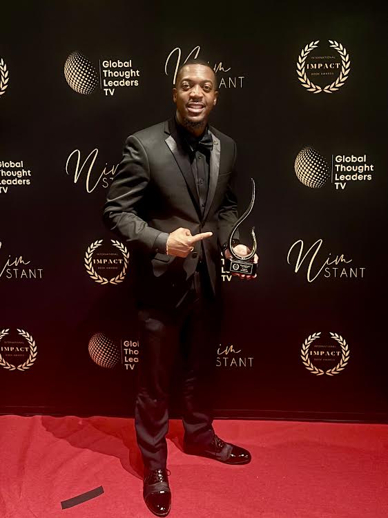 Man in a suit holding an award, standing on a red carpet with a black backdrop featuring logos and text.