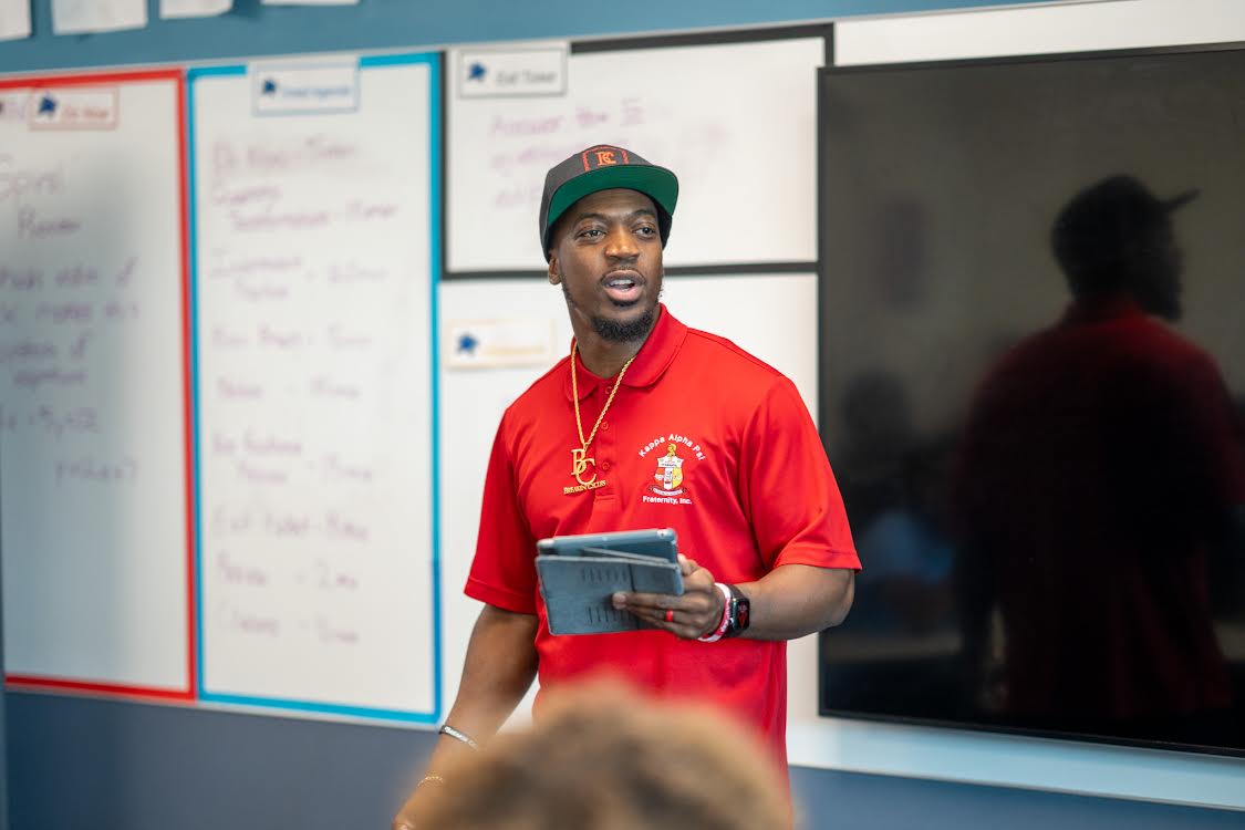 Young man in red shirt and cap holding a tablet, standing in front of a whiteboard and a black screen.