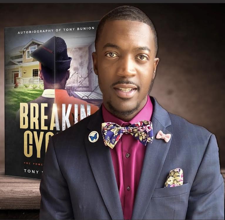 Man in a suit with a colorful bow tie and pocket square, standing in front of a book cover, smiling.
