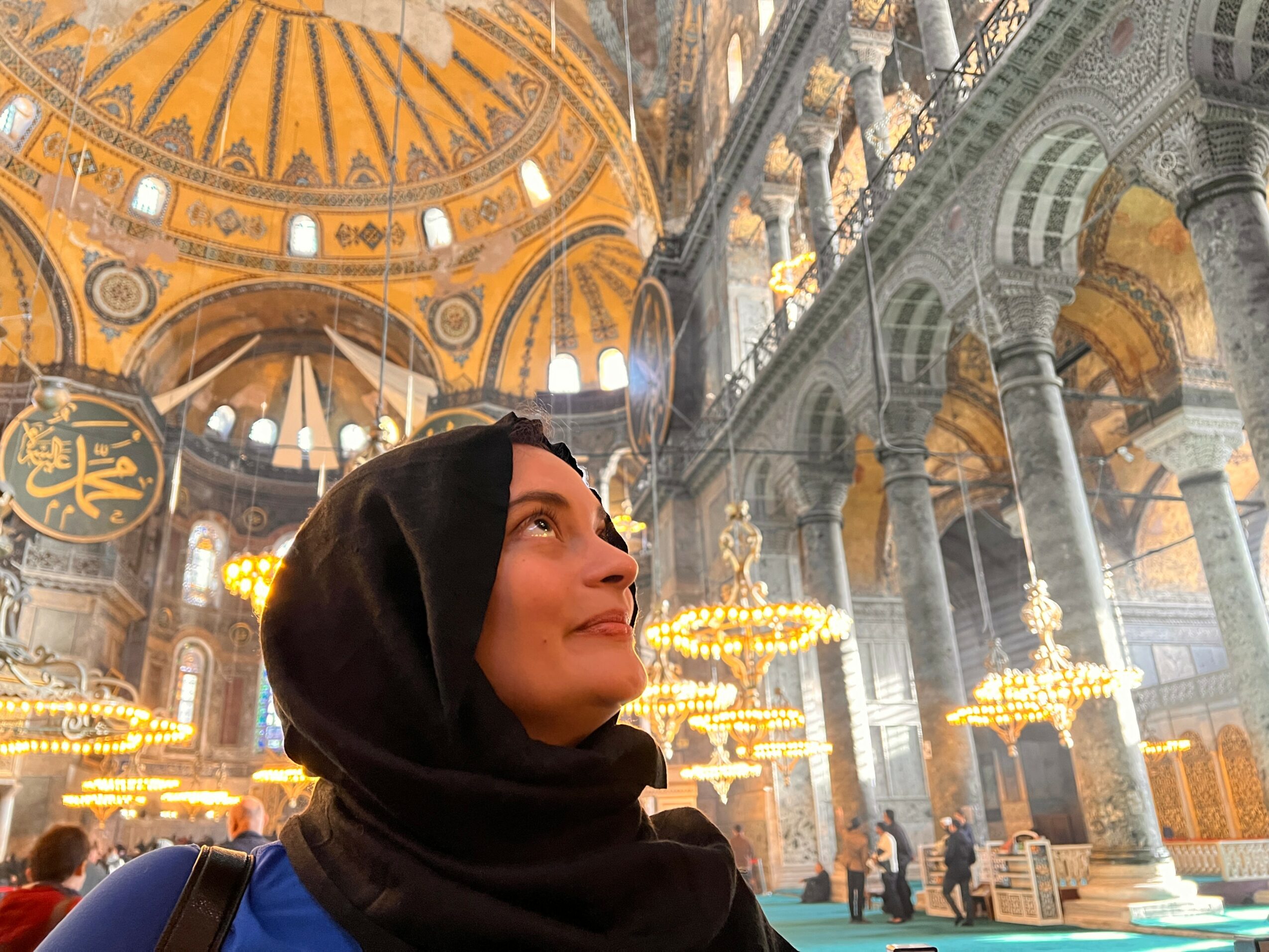 Woman wearing a black headscarf inside a grand, ornate mosque with golden domes and chandeliers.