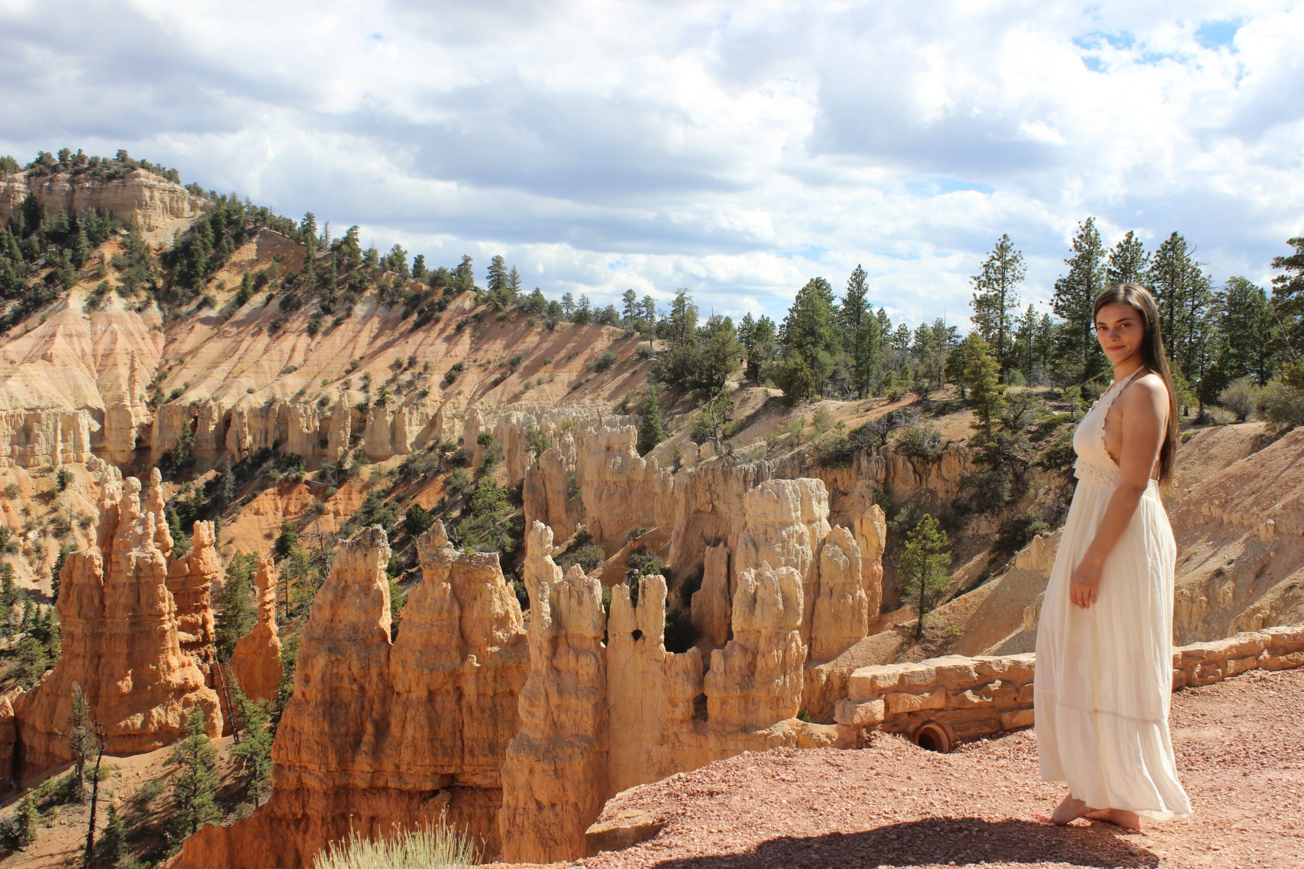 Woman in a white dress standing on rocky terrain with hoodoos and trees in the background under a partly cloudy sky.