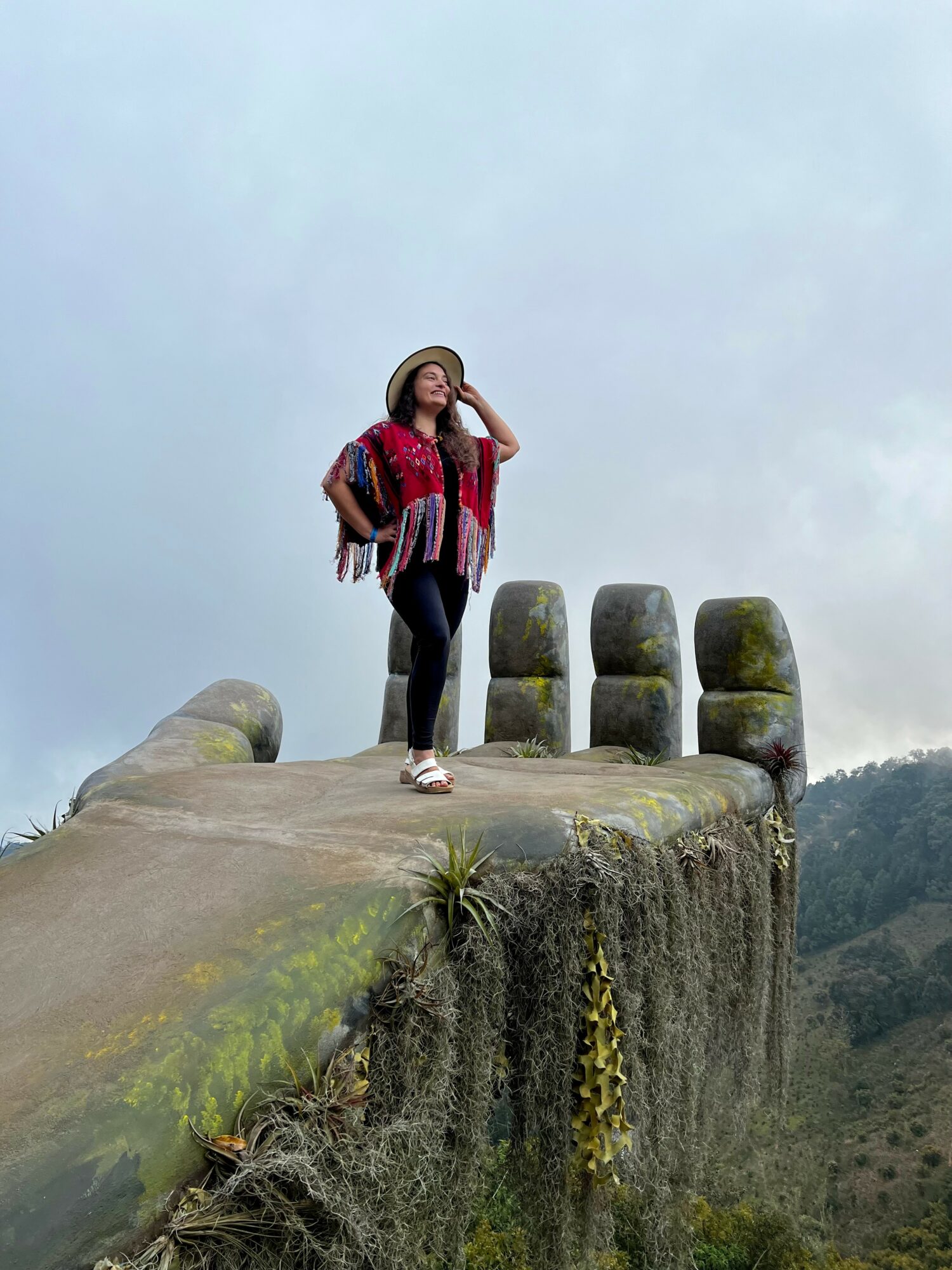 Woman standing on ancient stone structure with columns, wearing a hat and colorful clothing, overlooking a landscape.