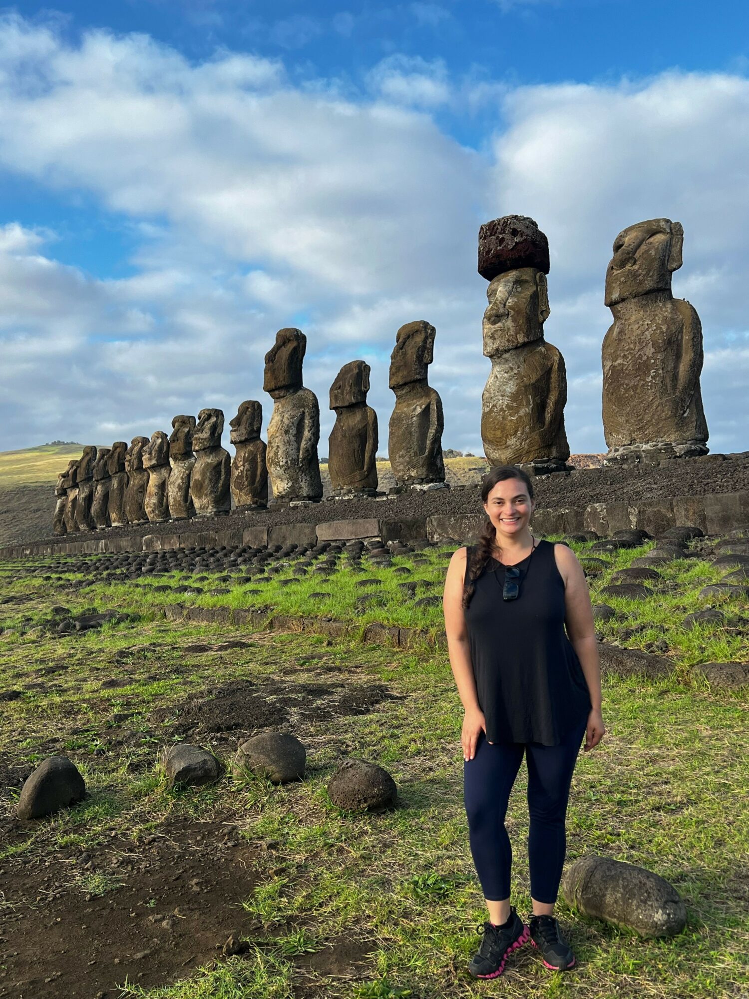 Person standing outdoors near stone statues on grassy ground with a cloudy sky background.